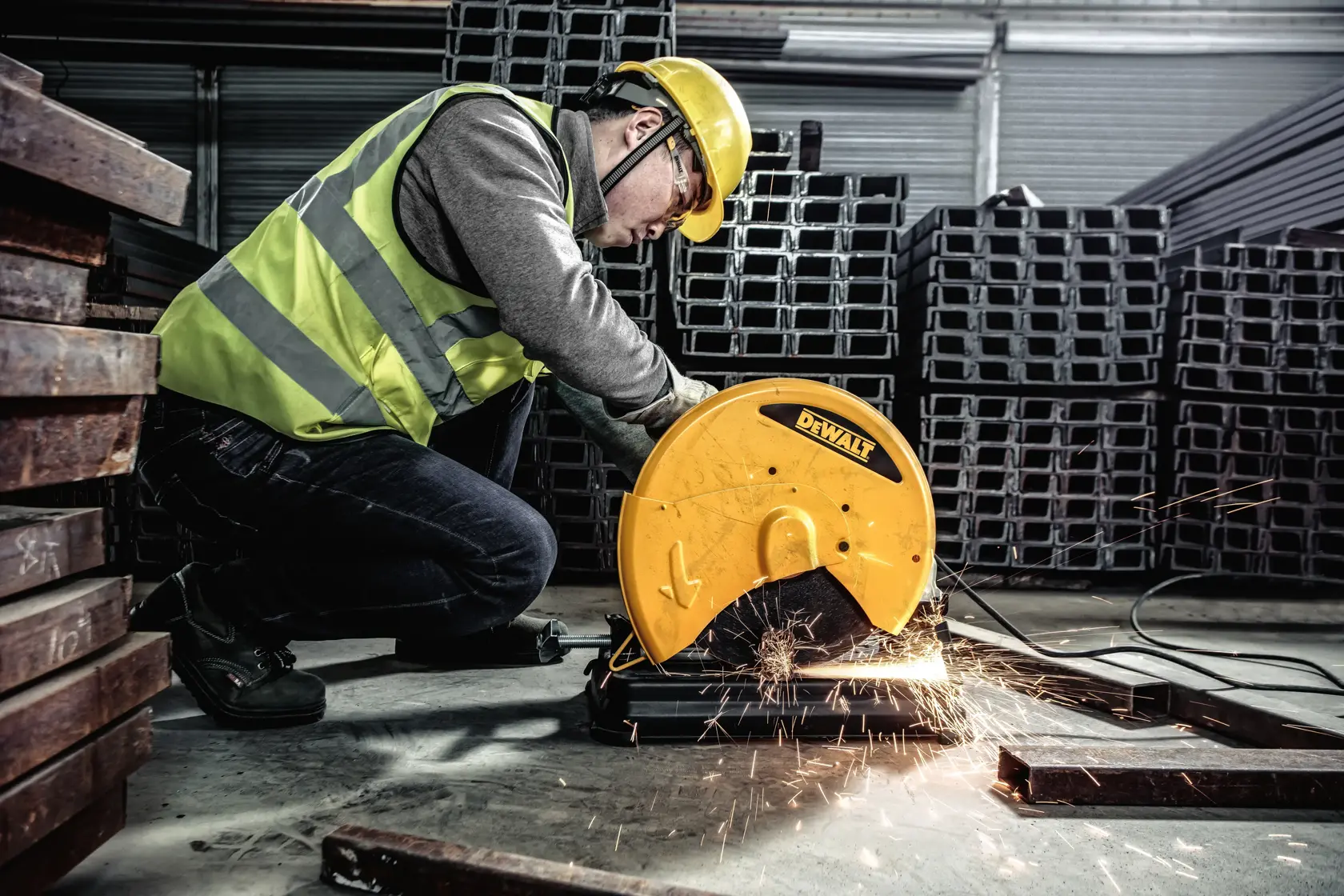 A worker wearing a yellow hard hat and reflective vest uses a yellow power saw to cut metal bars on the floor inside a large industrial warehouse. Sparks are visible from the cutting process.