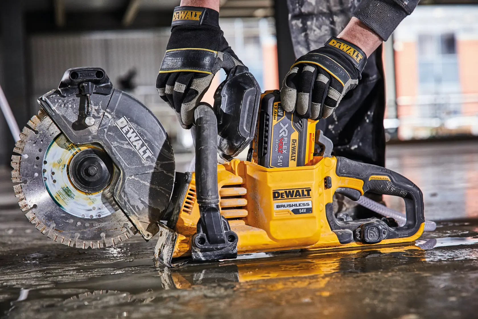 Eine Person mit Schutzhandschuhen bedient eine DEWALT 54V bürstenlose Akku-Trennsäge auf einem nassen Baustellenboden.
