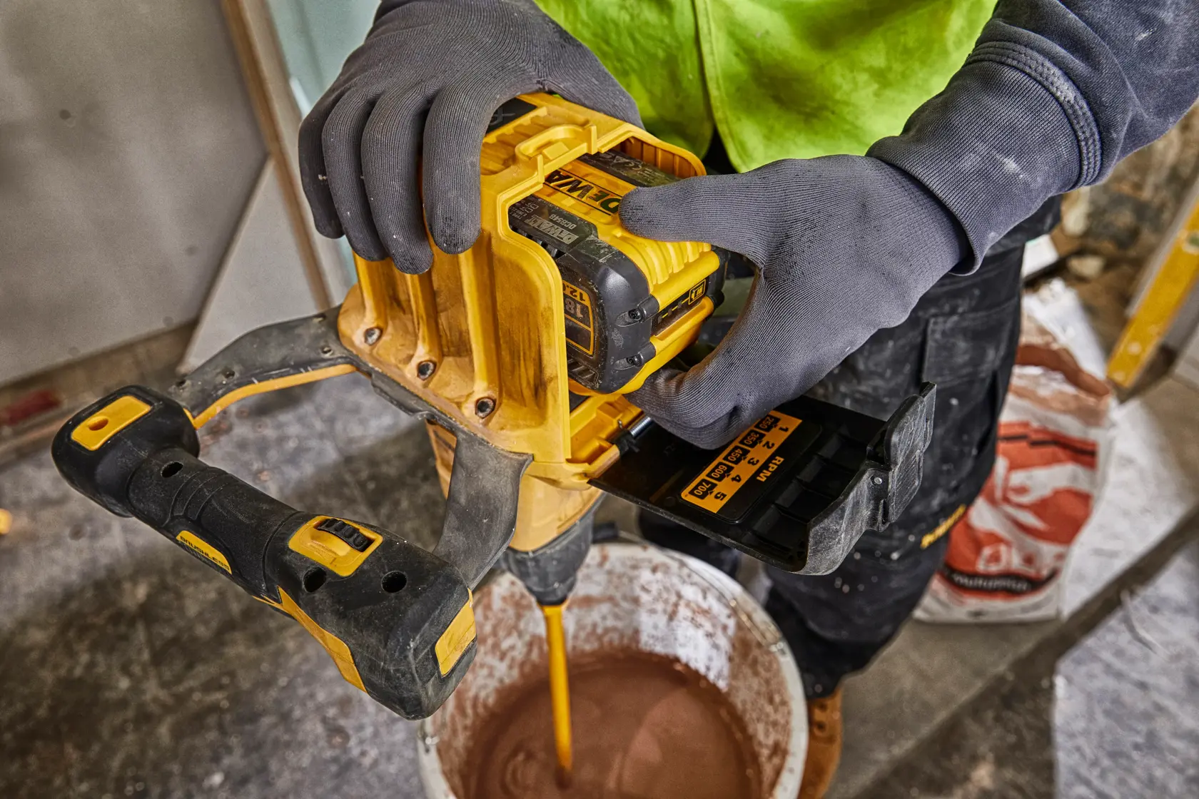 Eine Person mit Handschuhen verwendet einen gelben DEWALT-Akkumischer mit einem DEWALT DCB548-Akku, um Material in einem Eimer auf einer Baustelle zu mischen.