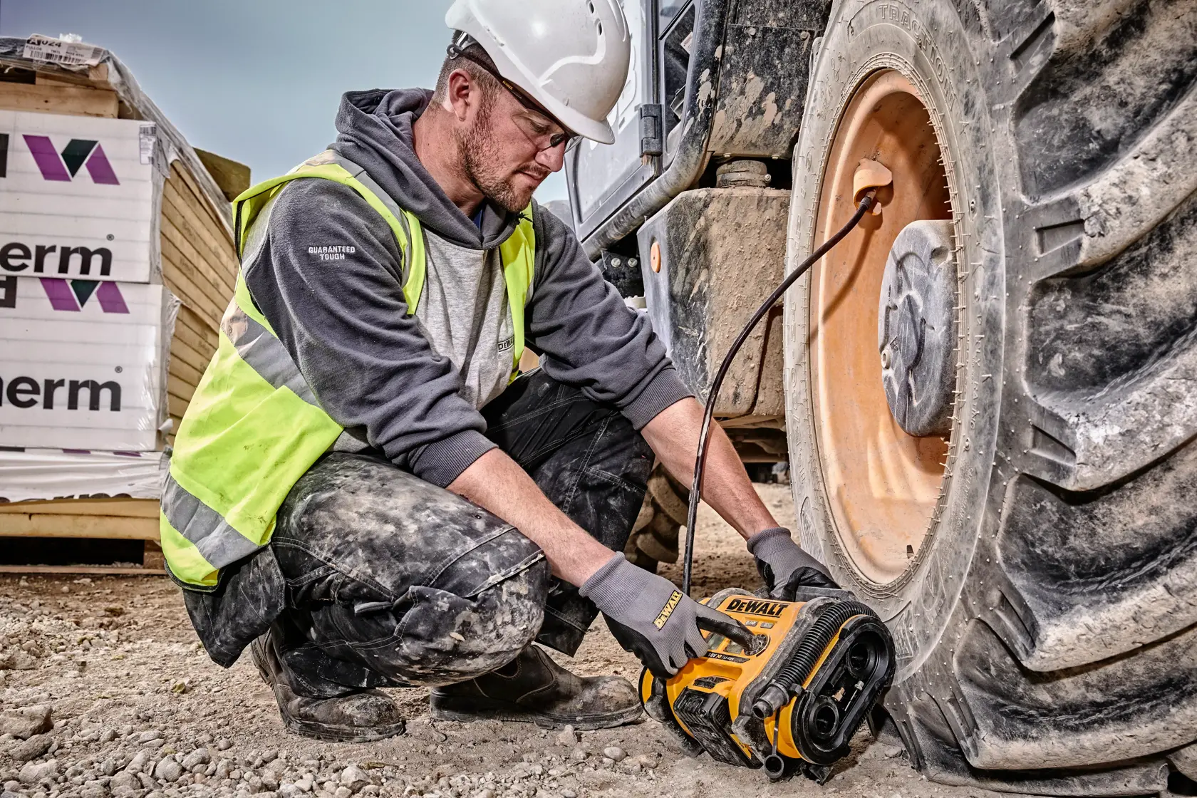 A person wearing a safety helmet and high-visibility vest is using a DEWALT DCC018N cordless air compressor to inflate the tire of a large construction vehicle on a construction site.