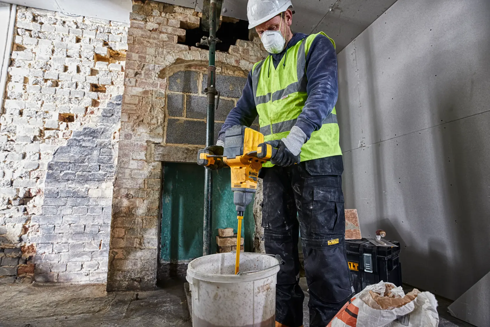 A construction worker wearing a high-visibility vest and hard hat is operating a yellow DEWALT DCD240N paddle mixer to mix materials in a white bucket inside a partially renovated room.