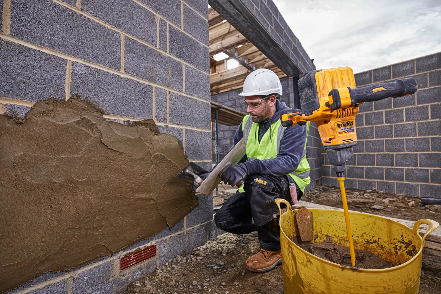 A construction worker wearing safety gear applies mortar to a brick wall using a trowel. Next to the worker, a yellow DEWALT DCD240N mixer is attached to a mixing paddle in a large yellow bucket with mortar. The scene is set at a construction site with unfinished brick walls.
