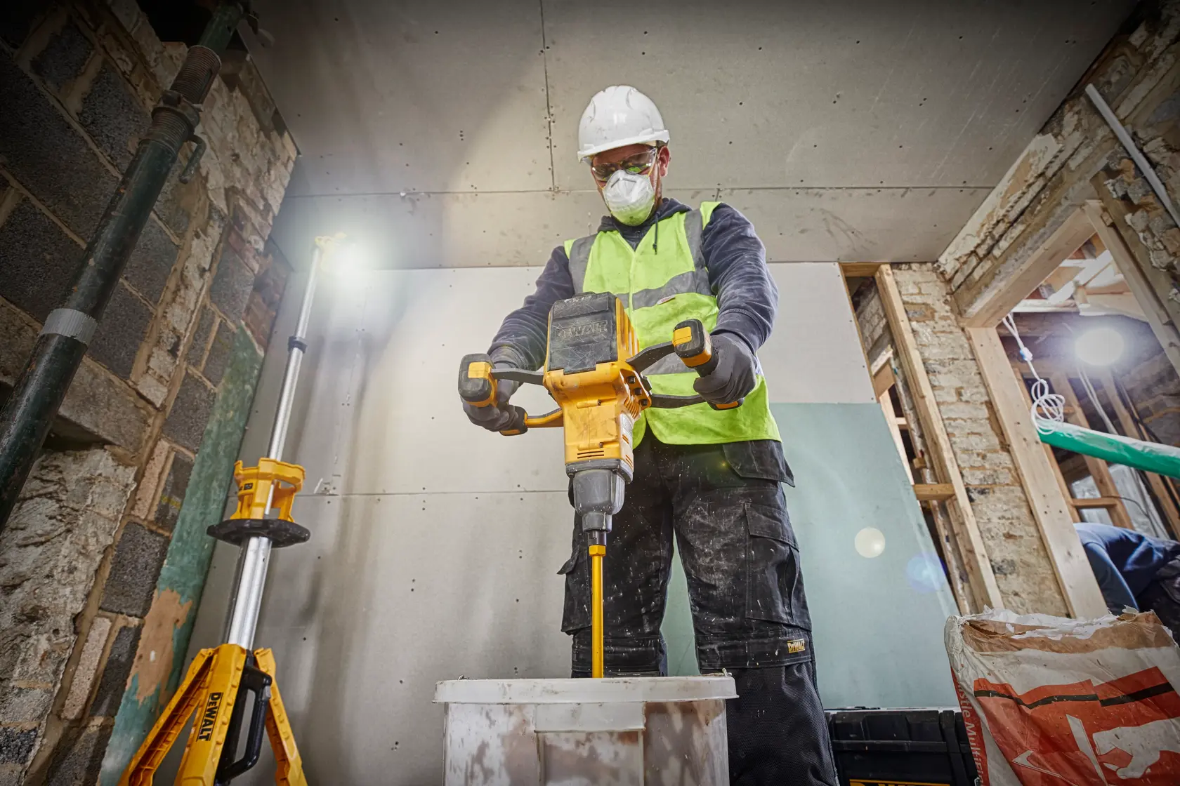 A construction worker wearing a hard hat and high-visibility vest uses a DEWALT DCD240X2 power mixer to mix material in a bucket, inside an unfinished building.