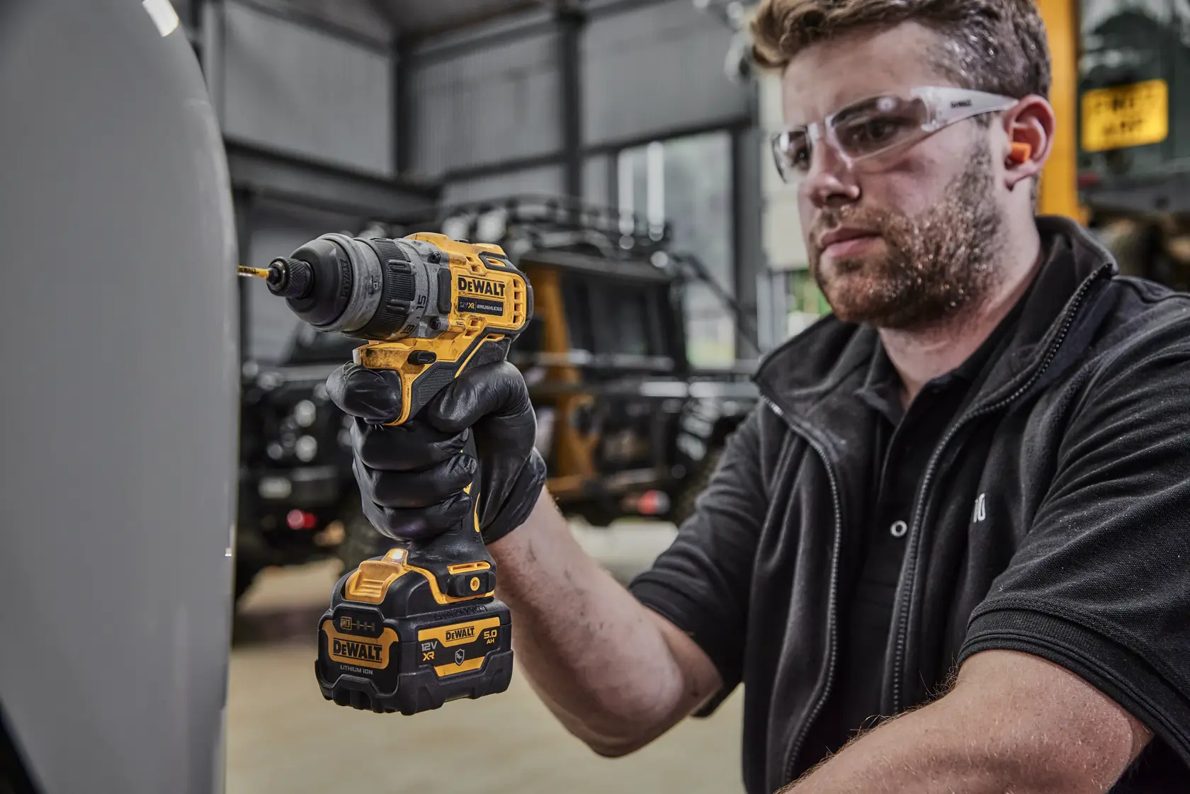 A person wearing black gloves is using a yellow and black DEWALT cordless drill with a battery attached indoors. The face of the person is blurred for privacy.