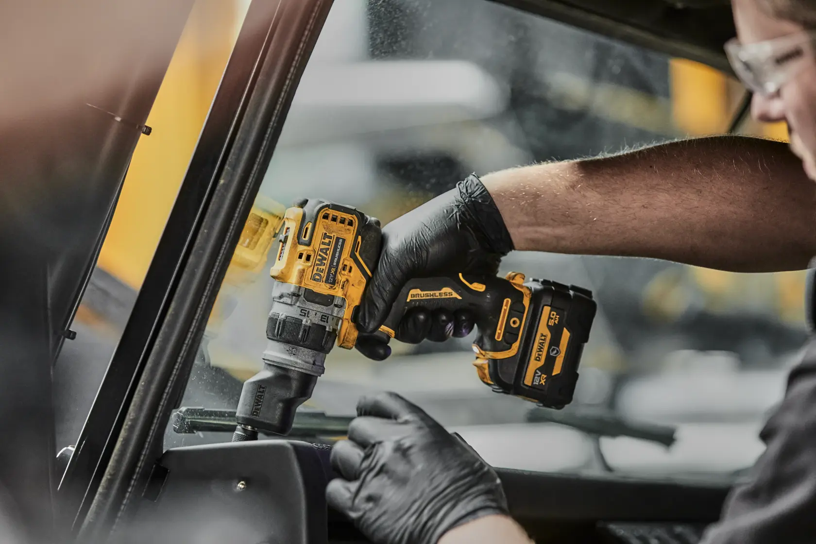 A person wearing black gloves uses a yellow and black DEWALT cordless drill with a brushless motor to work inside a vehicle near a window.
