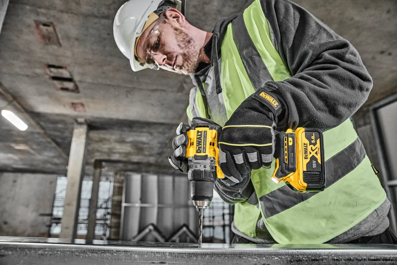 Une personne portant un équipement de sécurité, notamment des gants, un casque et un gilet haute visibilité, utilise une perceuse sans fil DEWALT DCD800P2T pour travailler sur une surface métallique dans un environnement industriel.