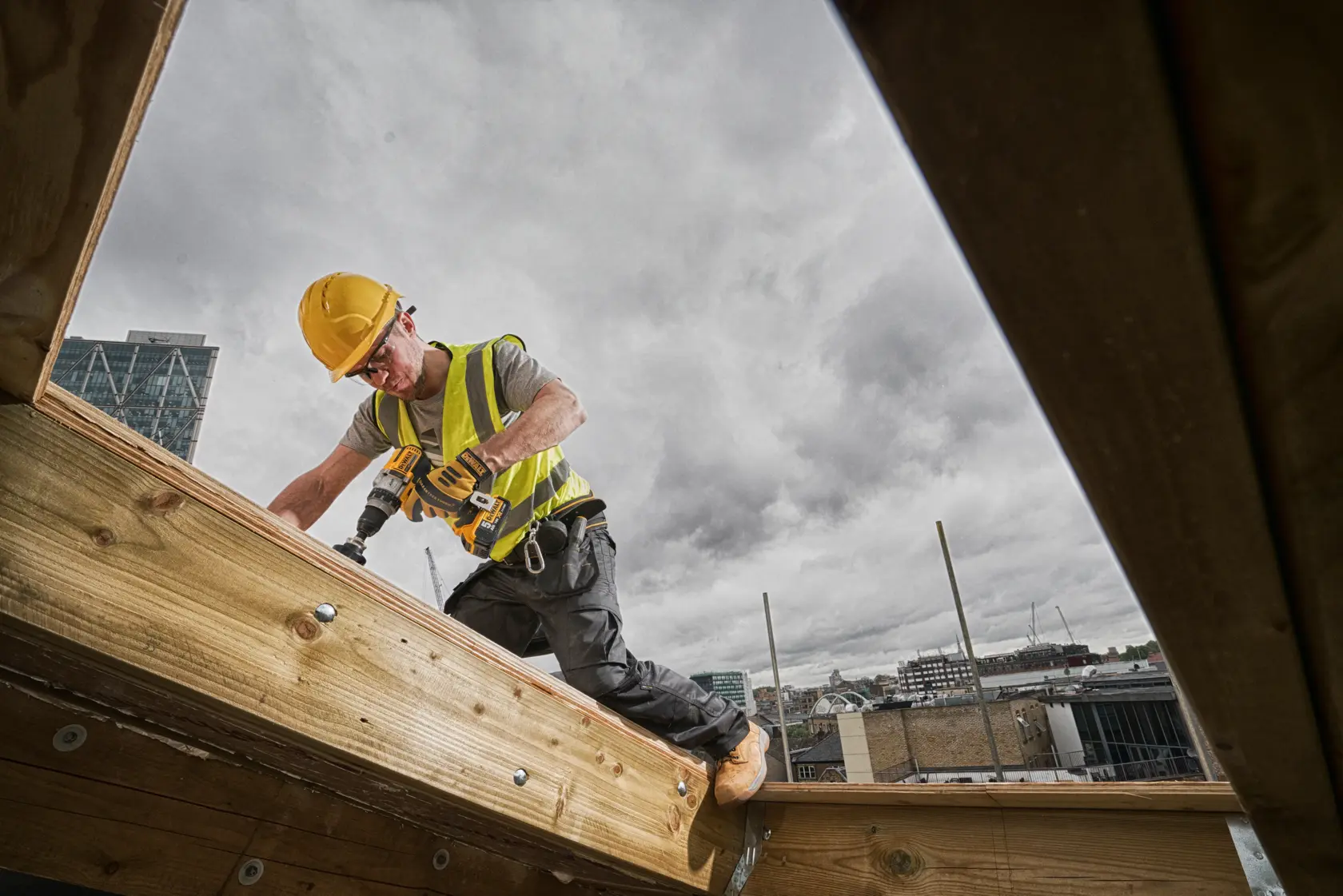 Ein Bauarbeiter mit gelbem Schutzhelm und Warnweste benutzt eine kabellose DEWALT-Bohrmaschine, während er im Freien auf einem Holzbalken arbeitet. Im Hintergrund sind Gebäude unter einem bewölkten Himmel sichtbar.