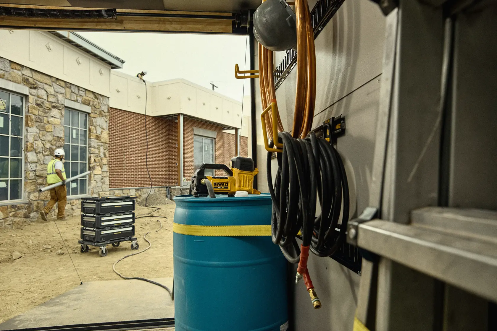 DEWALT device sits atop blue barrel in a worksite, coiled hoses and a worker can be seen outside near a stone building.