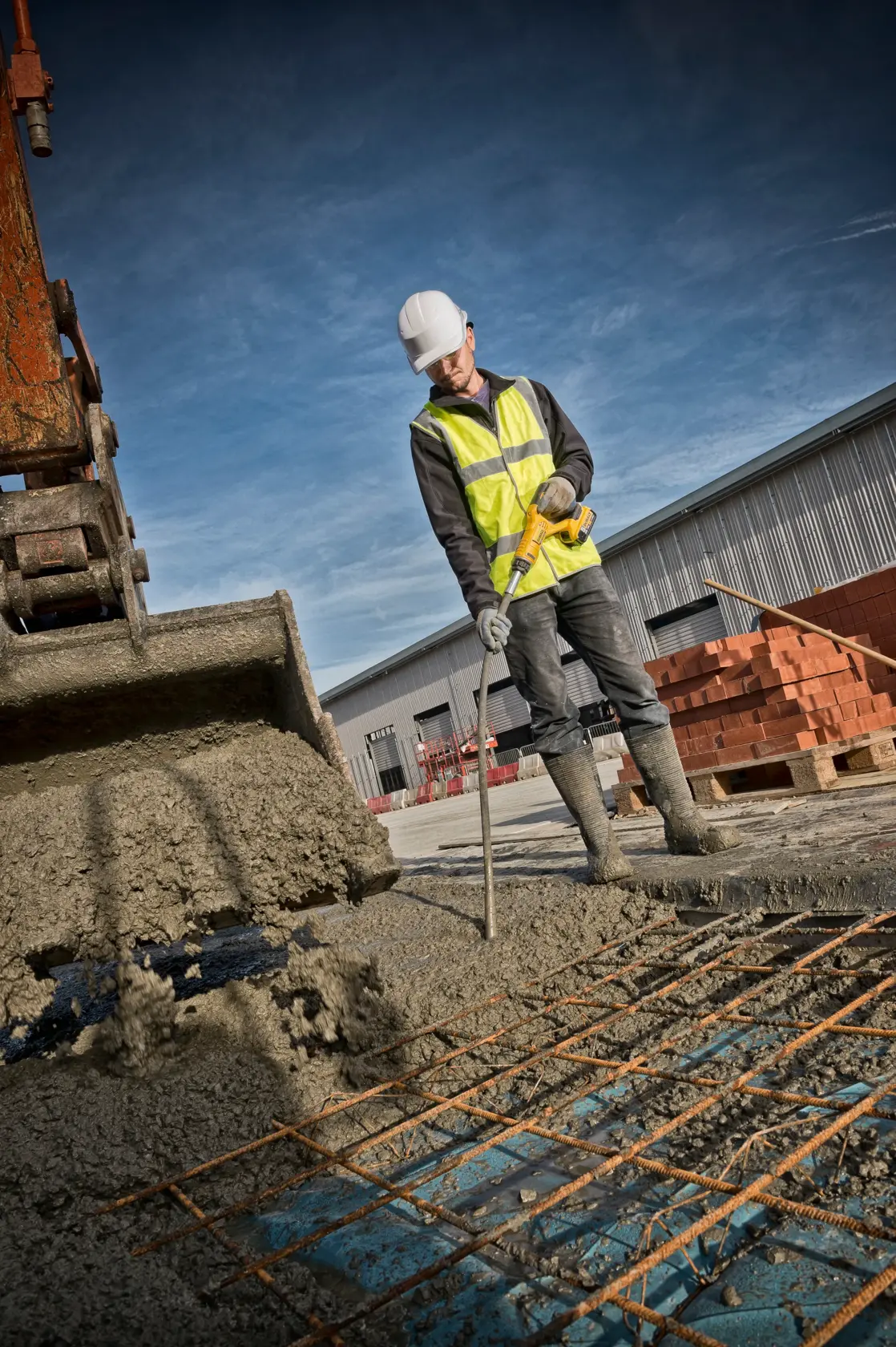 Ein Bauarbeiter mit Warnweste und Helm benutzt einen DEWALT Betonvibrator auf einer Baustelle, neben einem Bagger, der Beton gießt, und einem Stahlgitter.