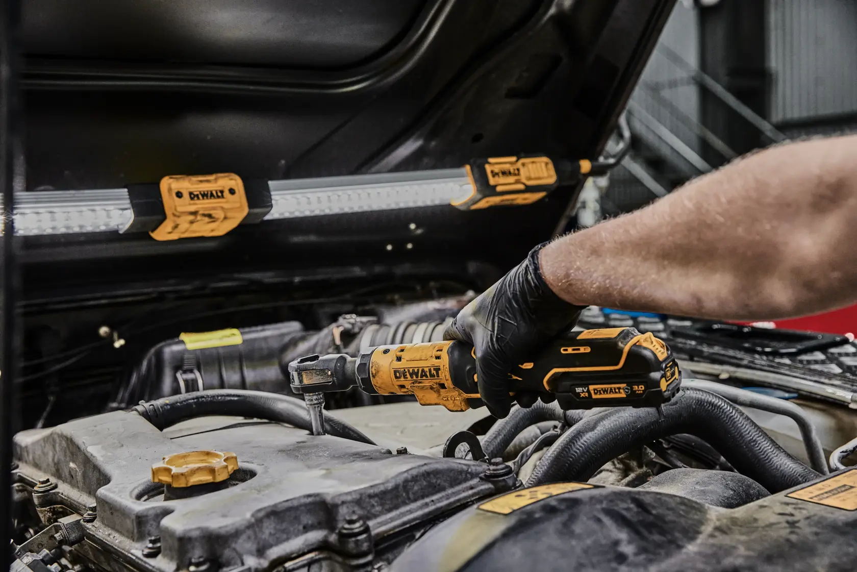 Una persona con guanti neri utilizza un attrezzo a cricchetto DEWALT senza fili su un motore di automobile, con una lampada da lavoro DEWALT fissata al cofano aperto.