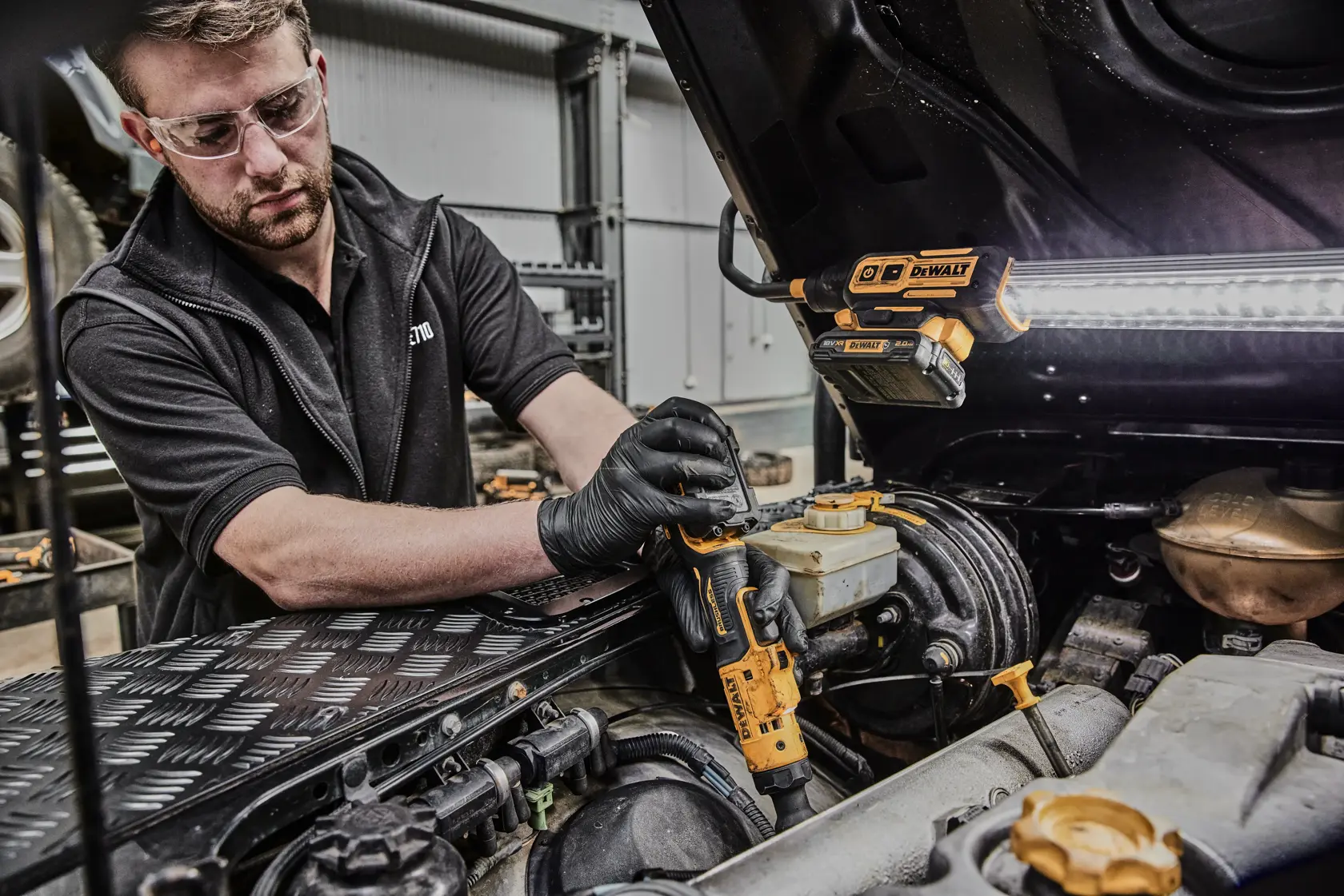 Una persona con guanti neri utilizza un avvitatore a cricchetto a batteria DEWALT DCF503N per lavorare sul motore di un'auto in un'officina. Una lampada da lavoro DEWALT è fissata al cofano dell'auto, illuminando l'area di lavoro.