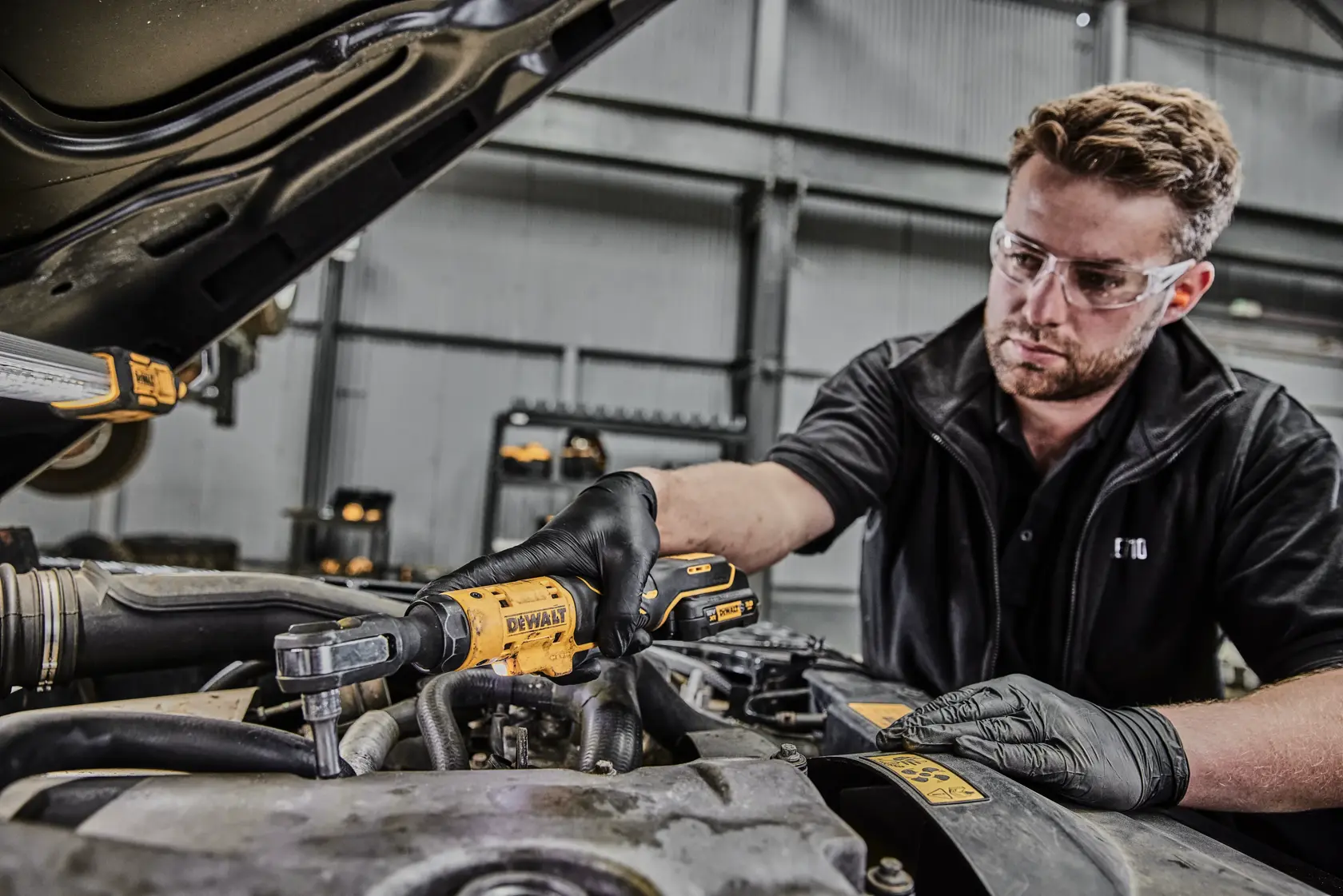 Eine Person mit schwarzen Handschuhen verwendet ein DEWALT DCF504L1G Akku-Ratschwerkzeug, um an einem Auto-Motor in einer Werkstatt zu arbeiten.