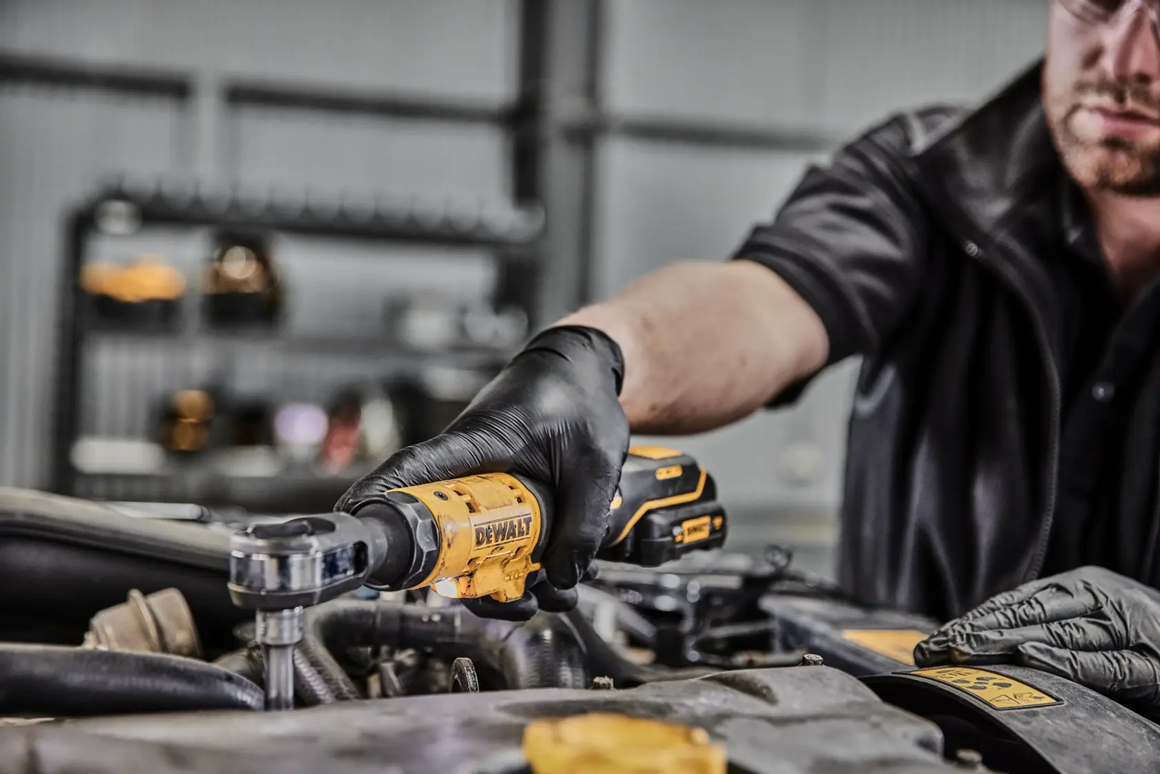 A person wearing black gloves uses a DEWALT DCF504N cordless ratchet tool to work on a car engine in a garage setting.