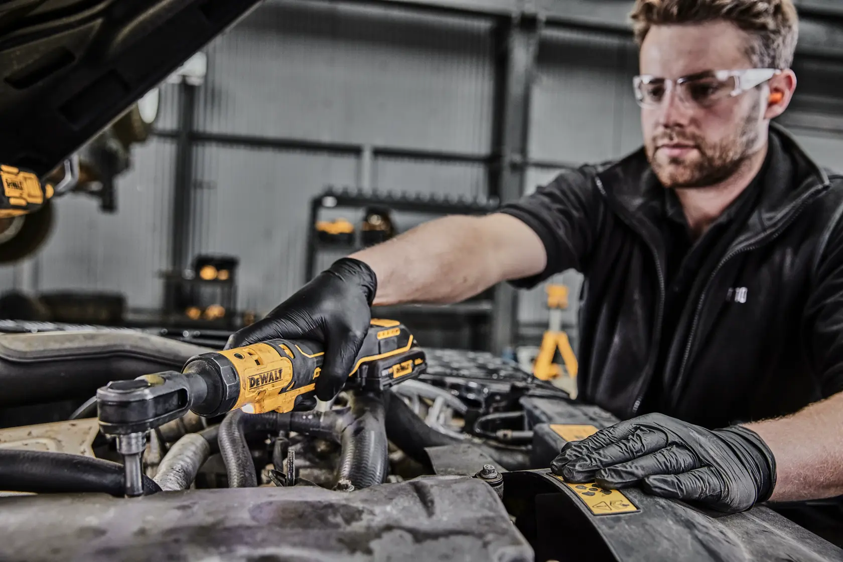 A person wearing black gloves is using a DEWALT cordless ratchet tool (DCF513N) to work on a car engine in a workshop. The