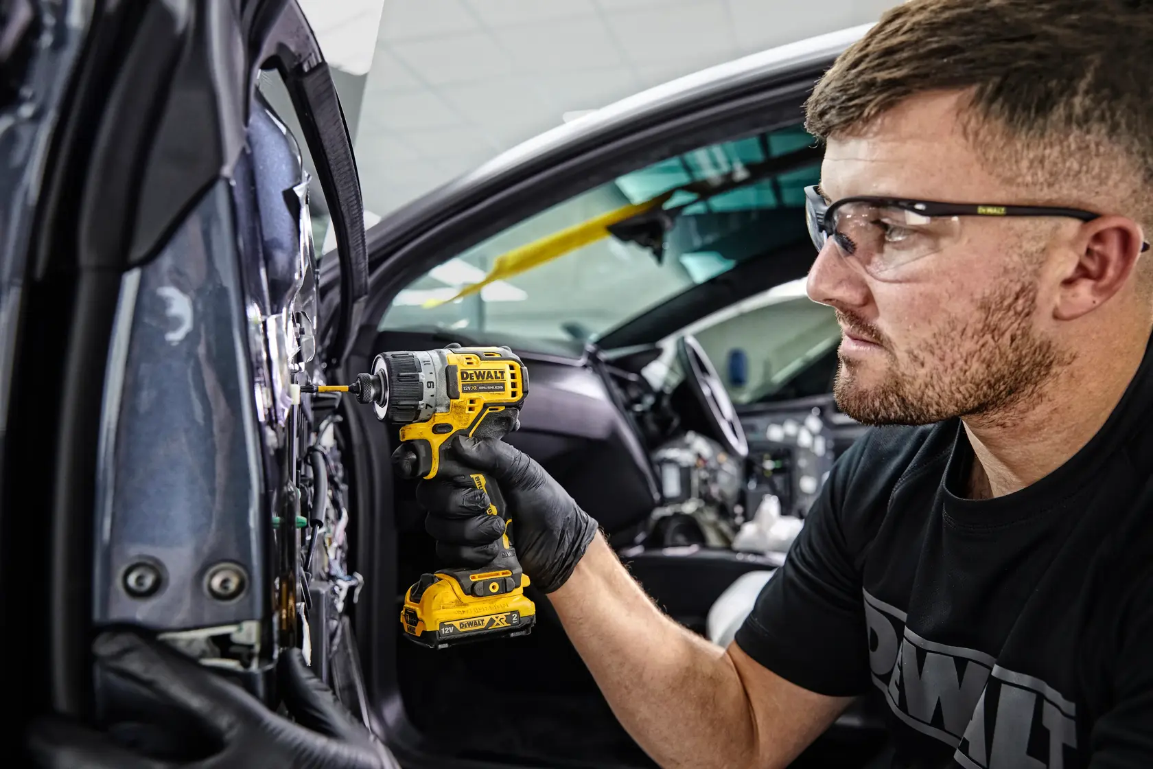 A person wearing black gloves uses a yellow DEWALT cordless screwdriver (DCF601D2) to work on the interior of a car door. The individual's face is blurred, and they are wearing a DEWALT-branded shirt.
