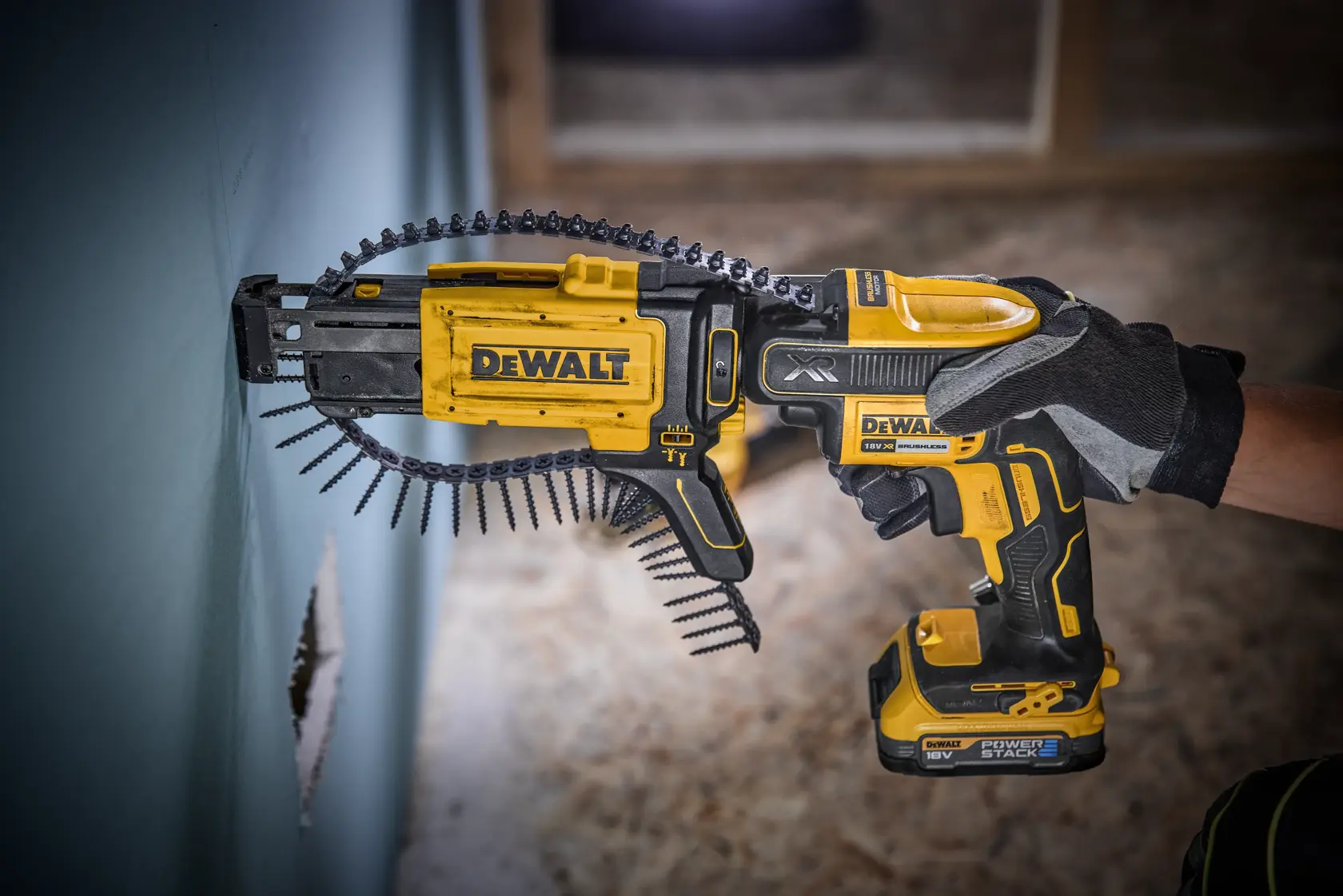 A DEWALT cordless drywall screw gun (DCF620E1K) being used to fasten screws into a wall. The tool is yellow and black, held by a gloved hand, with a strip of collated screws loaded in the gun.