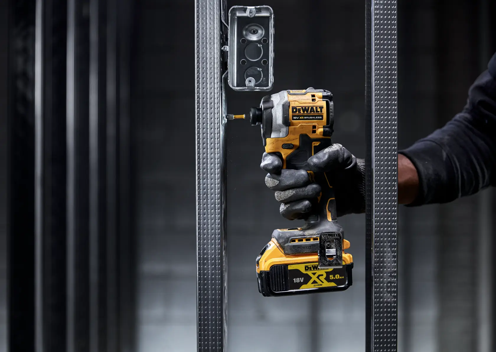 Mano enguantada usa un atornillador de impacto DEWALT en marco metálico cerca de caja eléctrica.