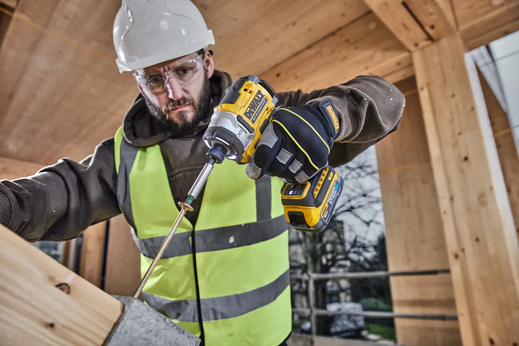 Worker uses a DEWALT DCF860E2T impact driver on wood, wearing safety gear at a construction site.