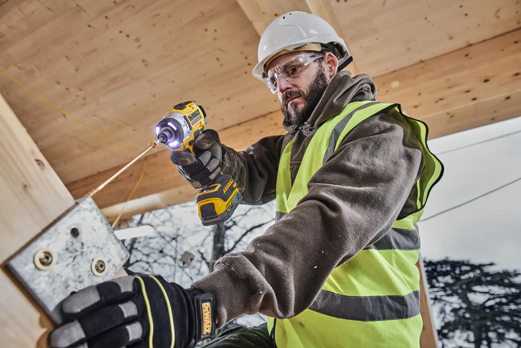 Worker uses DEWALT impact driver DCF860E2T on metal outdoors, wearing safety gear and gloves.