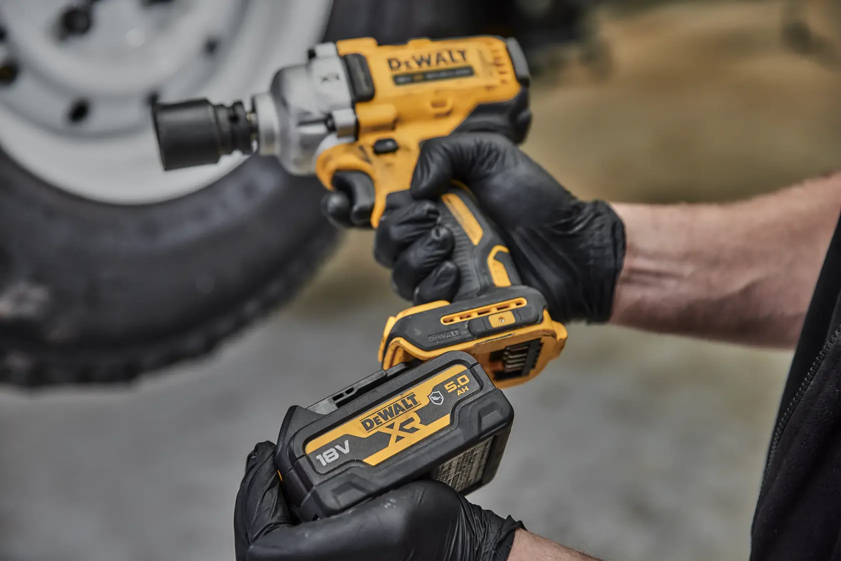 Behandschuhte Hände halten einen DEWALT Akku-Schlagschrauber und eine 18V Batterie vor einem Reifen.