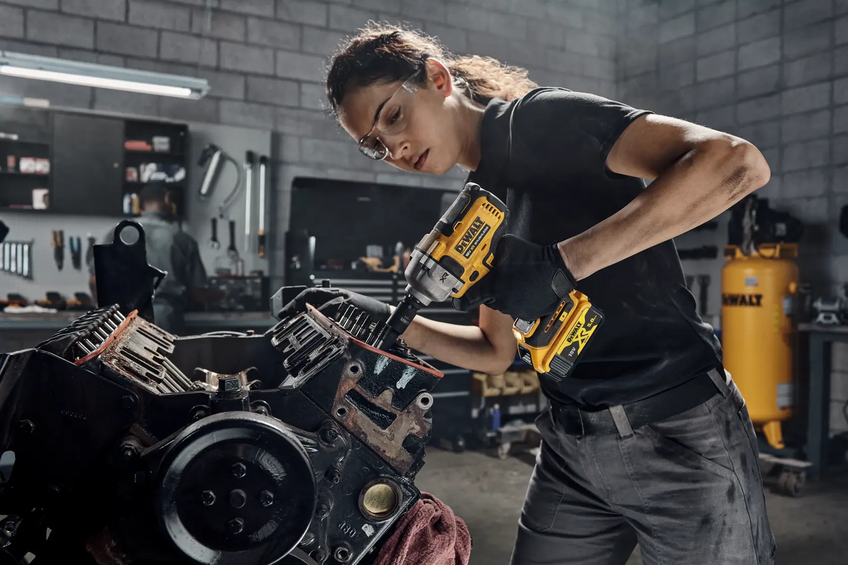 Person using a DEWALT DCF892P2T impact wrench on a motor in a workshop with industrial equipment.
