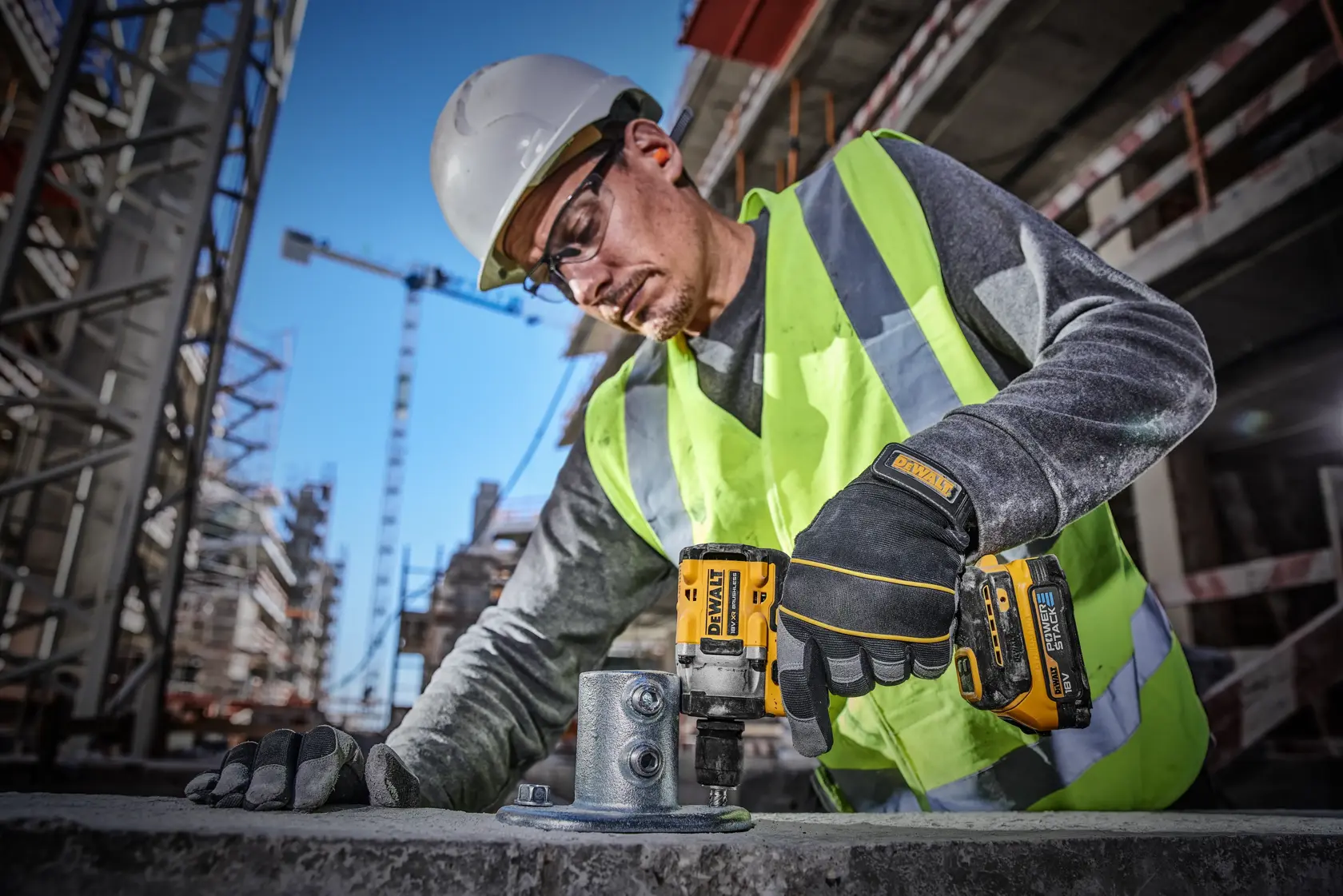 Un trabajador de la construcción con casco de seguridad, chaleco de alta visibilidad y guantes está usando una llave de impacto inalámbrica DEWALT DCF921E2T para asegurar pernos en una estructura metálica en un sitio de construcción concurrido.