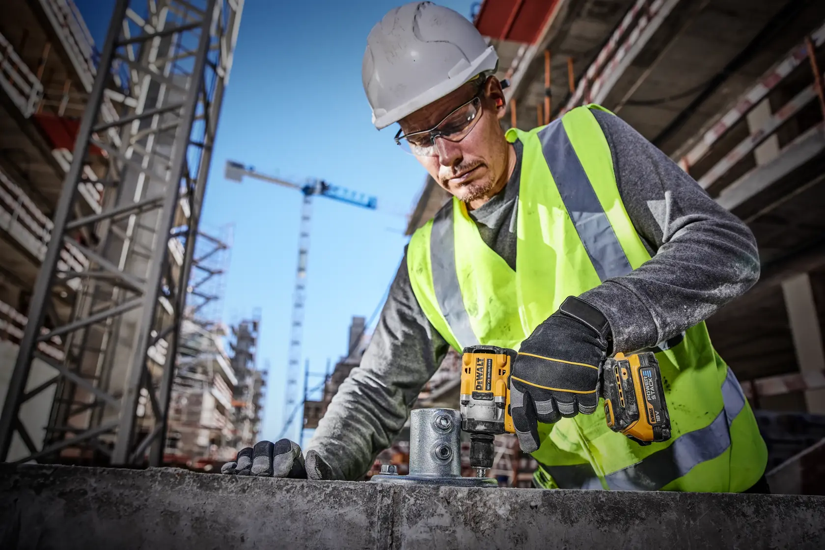 Un trabajador de la construcción con un chaleco de alta visibilidad y casco de seguridad utiliza una herramienta eléctrica DEWALT DCF921E2T en una obra. Se pueden ver varias grúas y edificios sin terminar en el fondo.