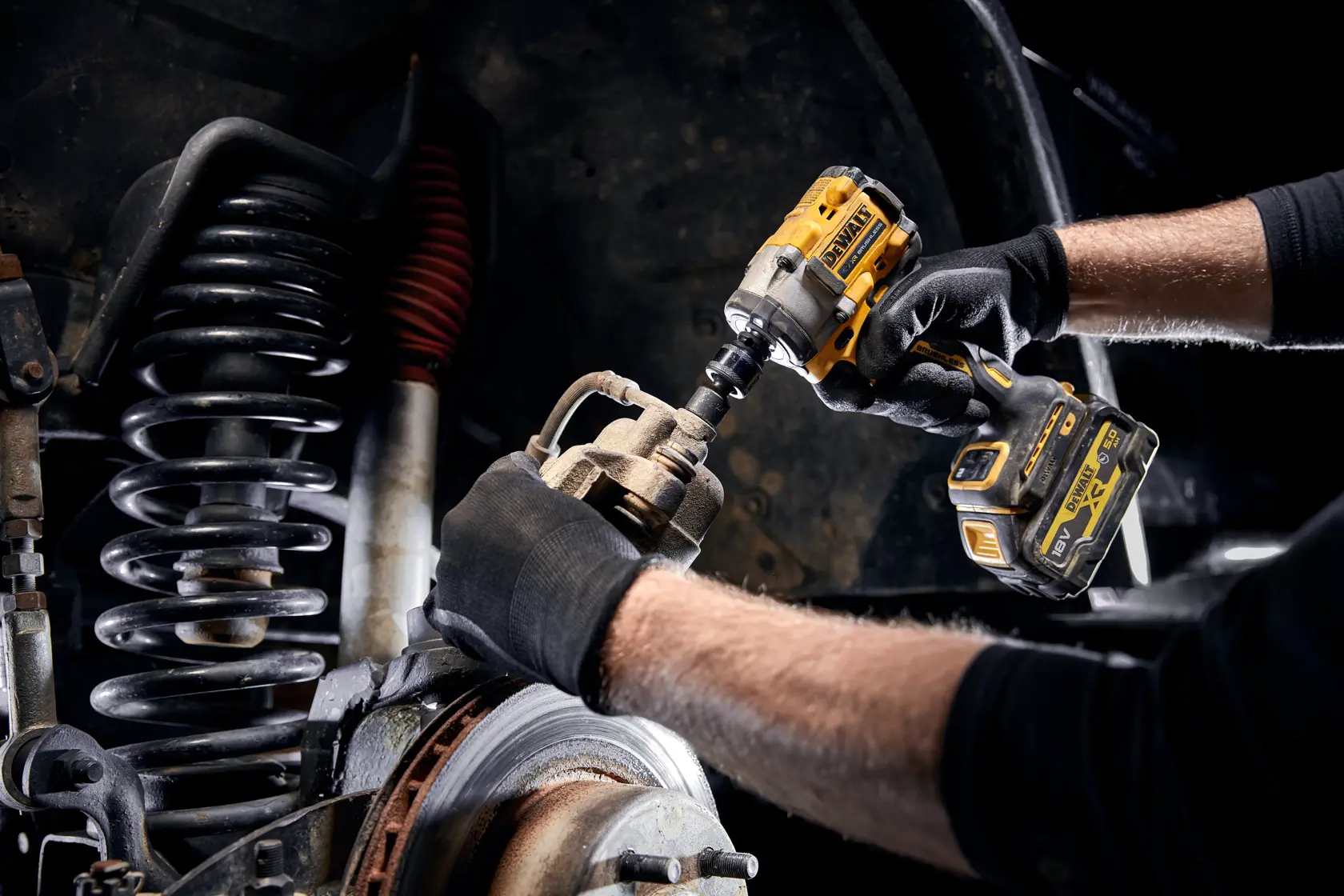 Eine Person mit schwarzen Handschuhen verwendet einen kabellosen DEWALT-Schlagschrauber, um an der Federung und den Bremskomponenten eines Fahrzeugs zu arbeiten.