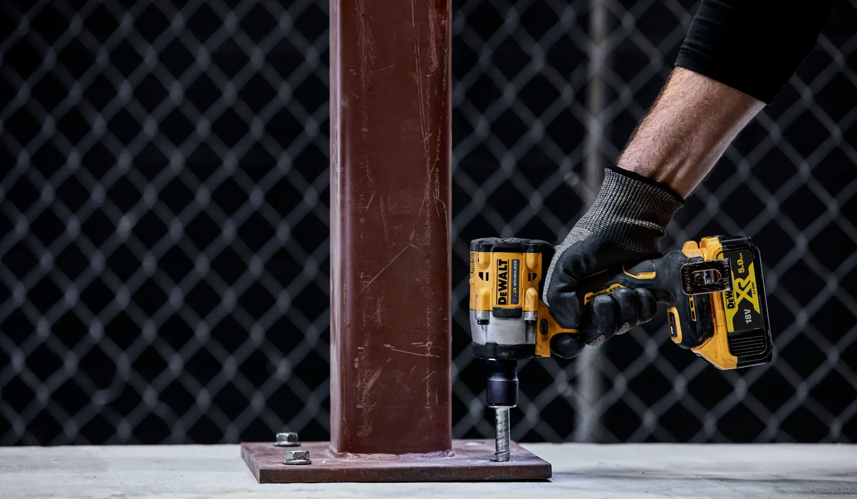 A person wearing a black glove uses a DEWALT cordless impact wrench to tighten a bolt on the base of a metal column, with a chain-link fence in the background.