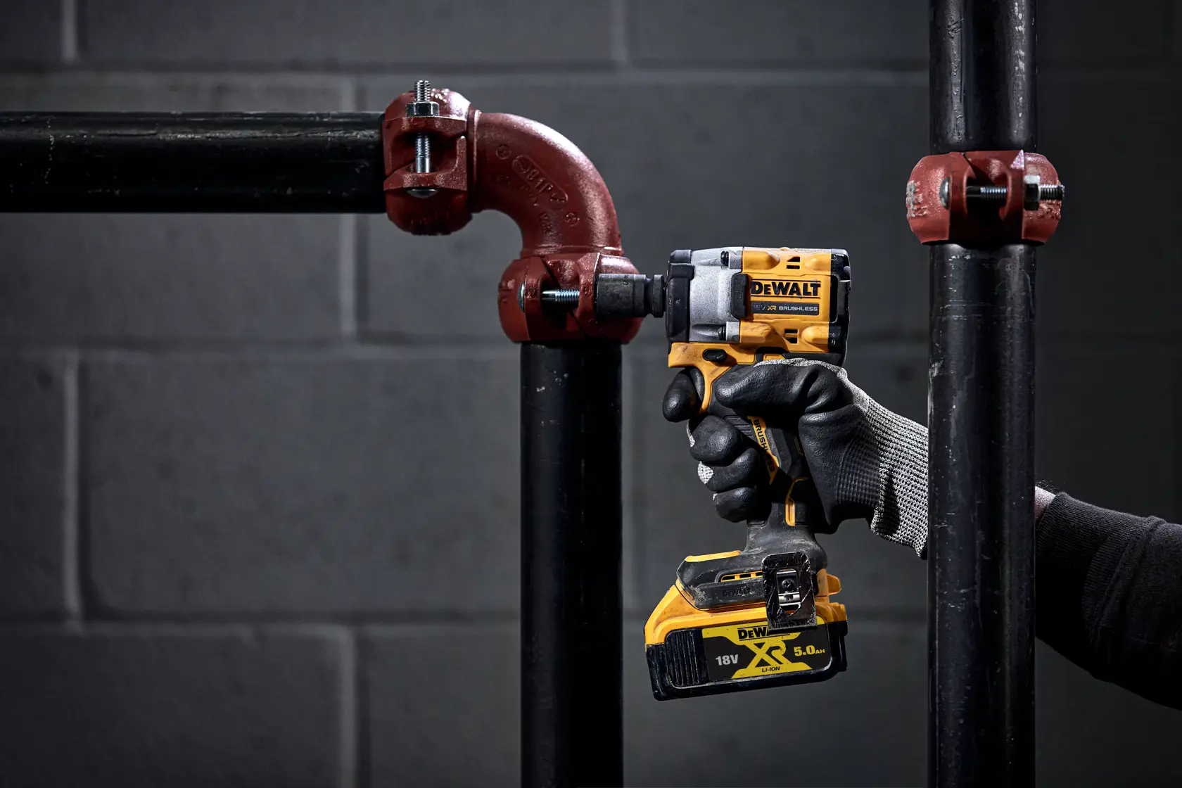 A person wearing gloves uses a DEWALT cordless impact wrench to fasten a bolt on a metal pipe fitting. The tool is yellow and black, and the scene is set against a dark, industrial background.