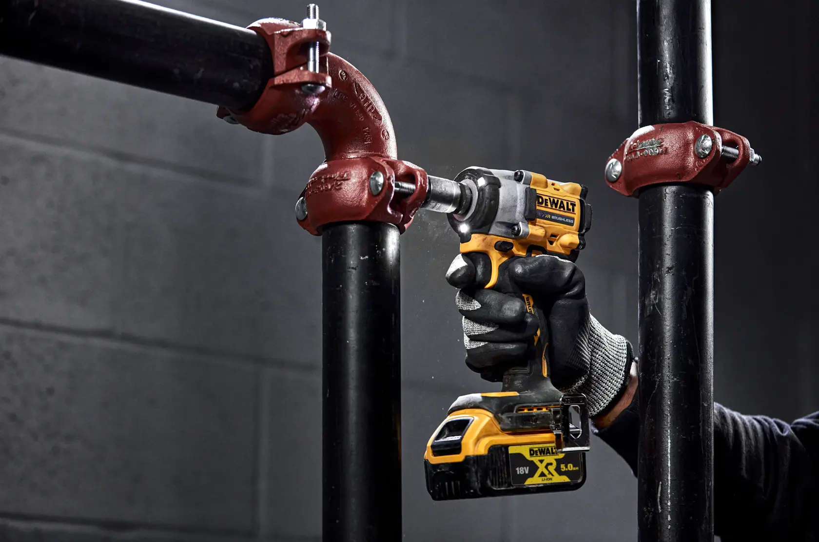 A person wearing work gloves uses a yellow and black DEWALT cordless impact wrench to tighten a bolt on a black metal pipe with a red fitting.