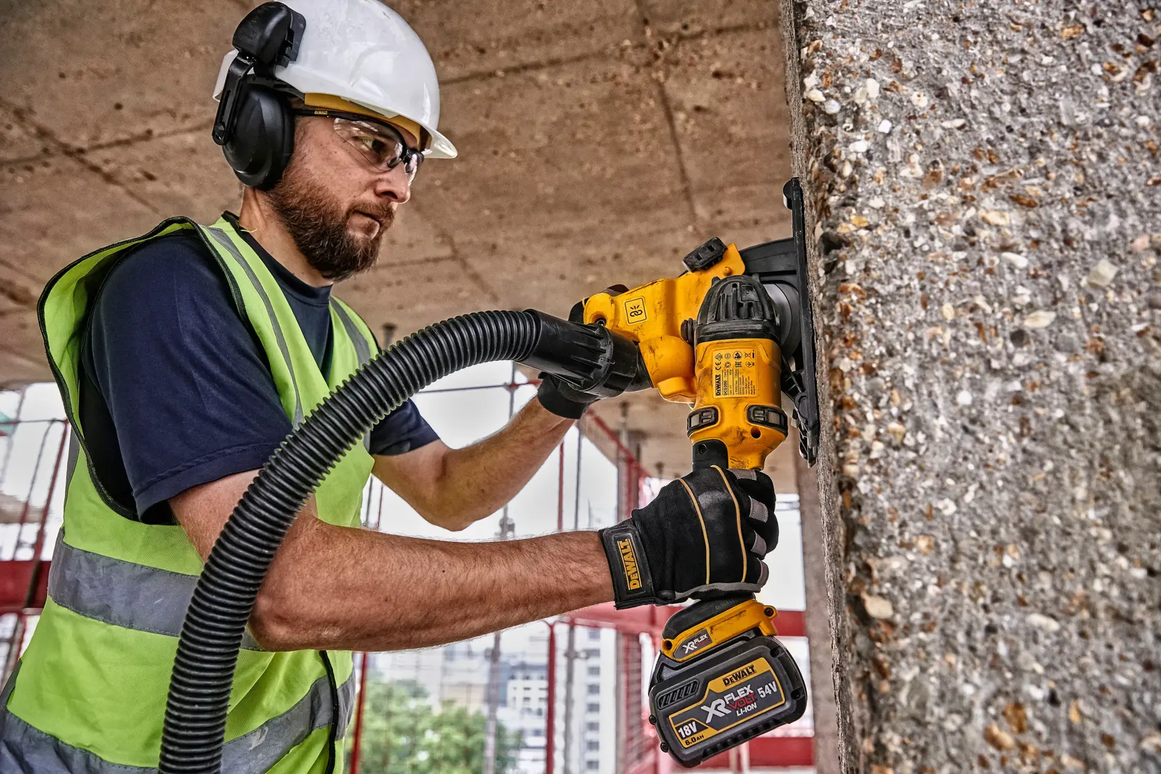 Un trabajador con casco de seguridad, protección auditiva y chaleco de alta visibilidad utiliza una herramienta eléctrica inalámbrica DEWALT DCG200T2 para pulir concreto en un sitio de construcción. El rostro del trabajador está difuminado para preservar la privacidad.