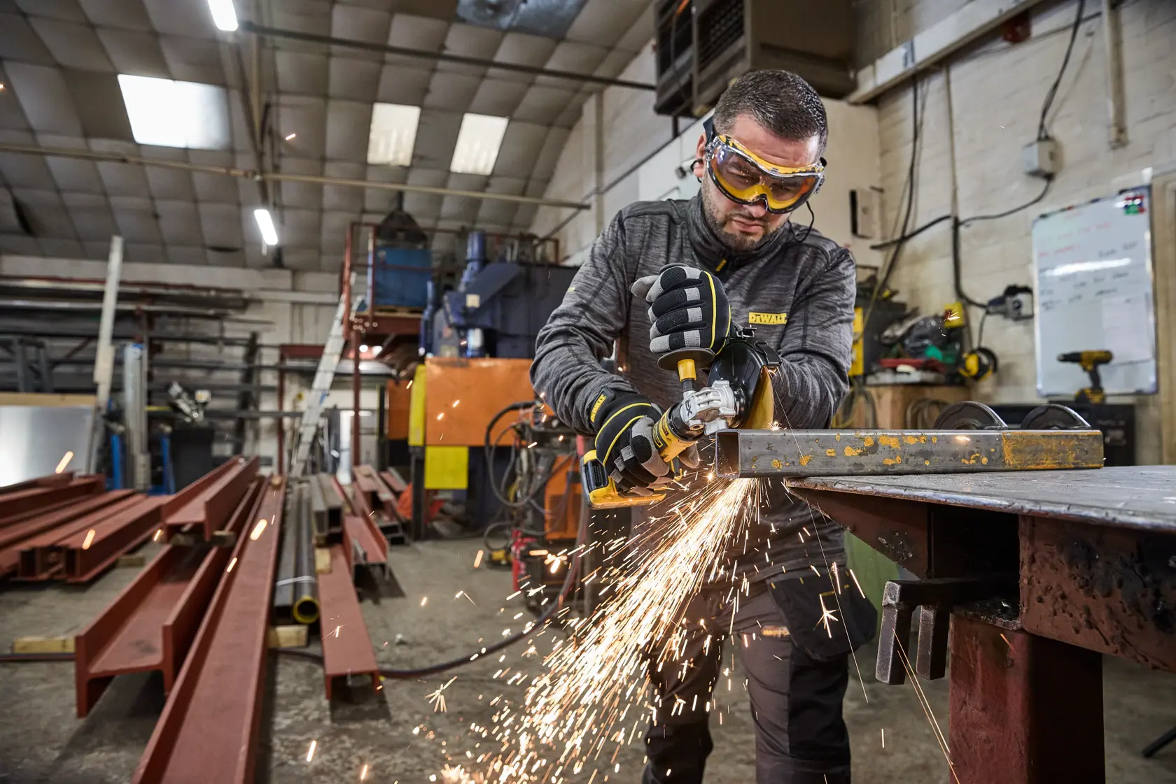 Person using a DEWALT angle grinder on metal in a workshop, producing sparks. Industrial equipment surrounds them.