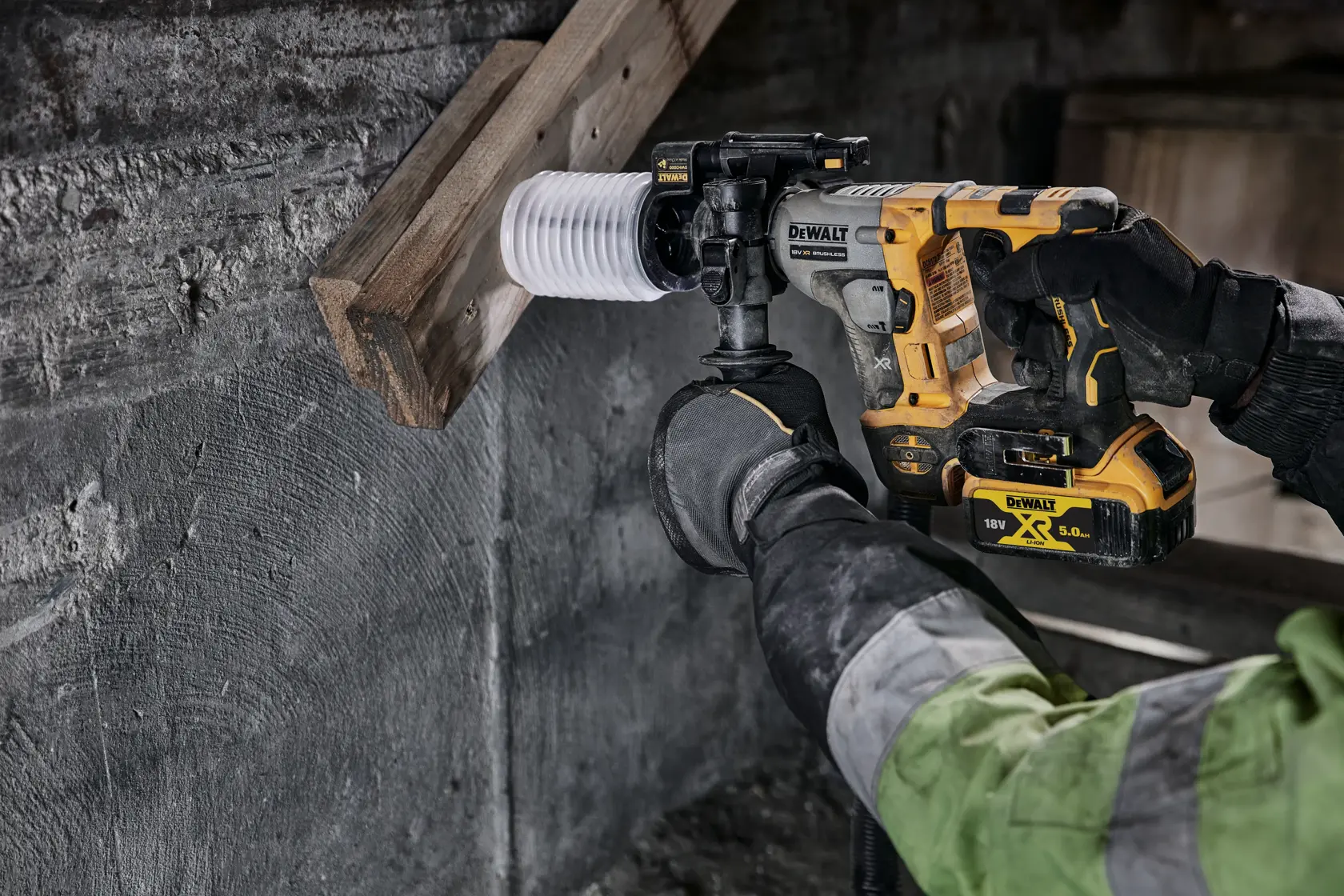 DEWALT cordless rotary hammer drilling into wood and concrete, operated by a gloved worker wearing a green jacket.