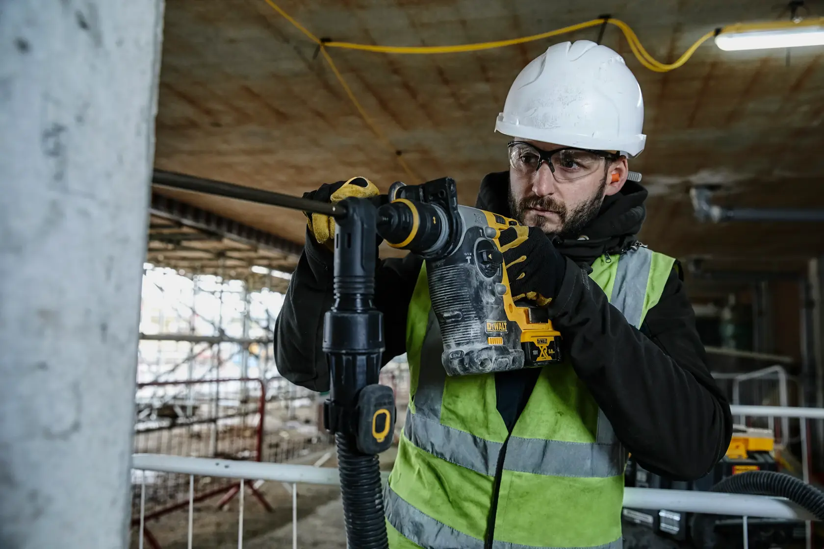 Worker using DEWALT DCH274N tool to drill into concrete at a construction site, wearing safety gear and vest.