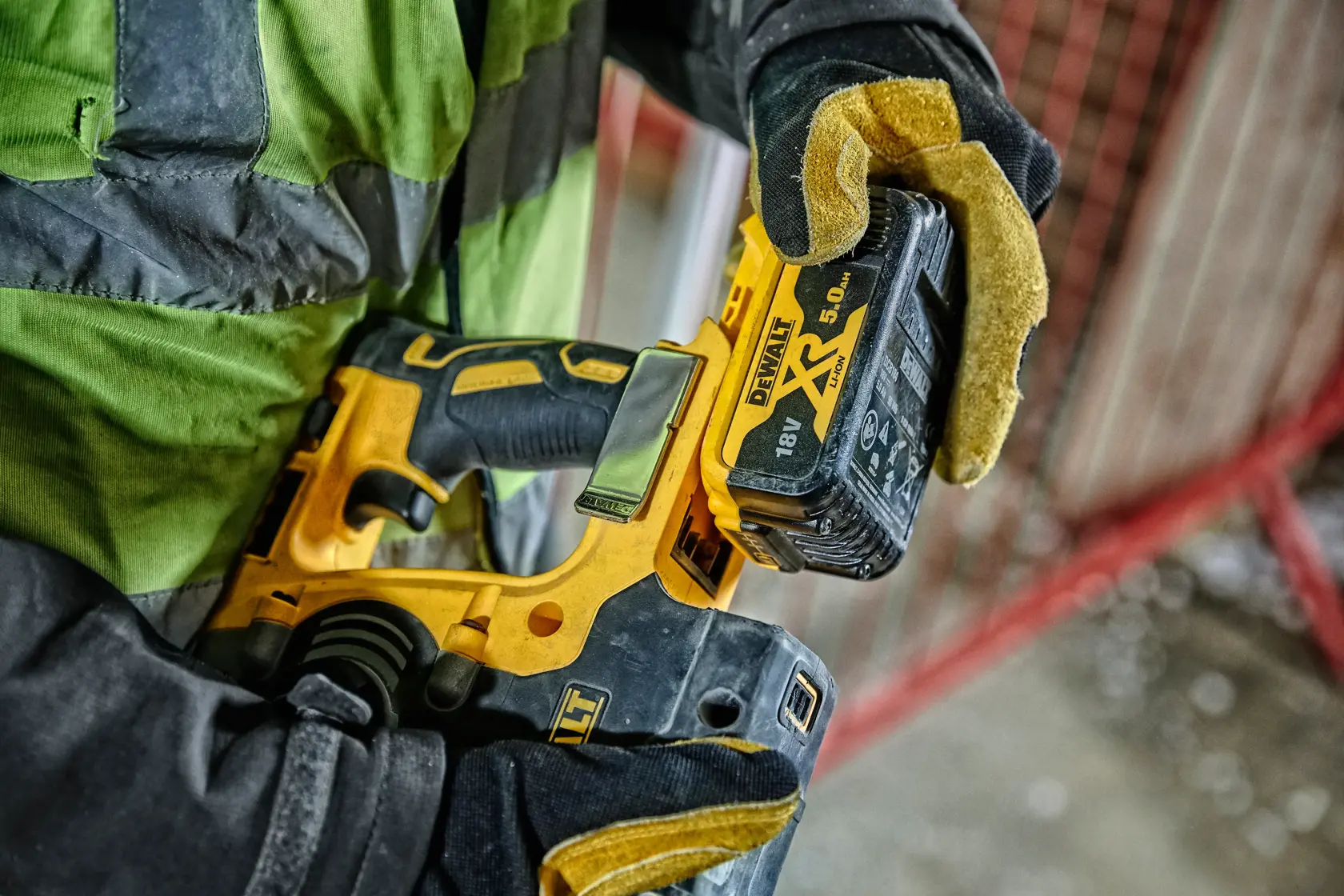 Gloved hand holds a DEWALT cordless power tool with an 18V XR lithium battery at a construction site.