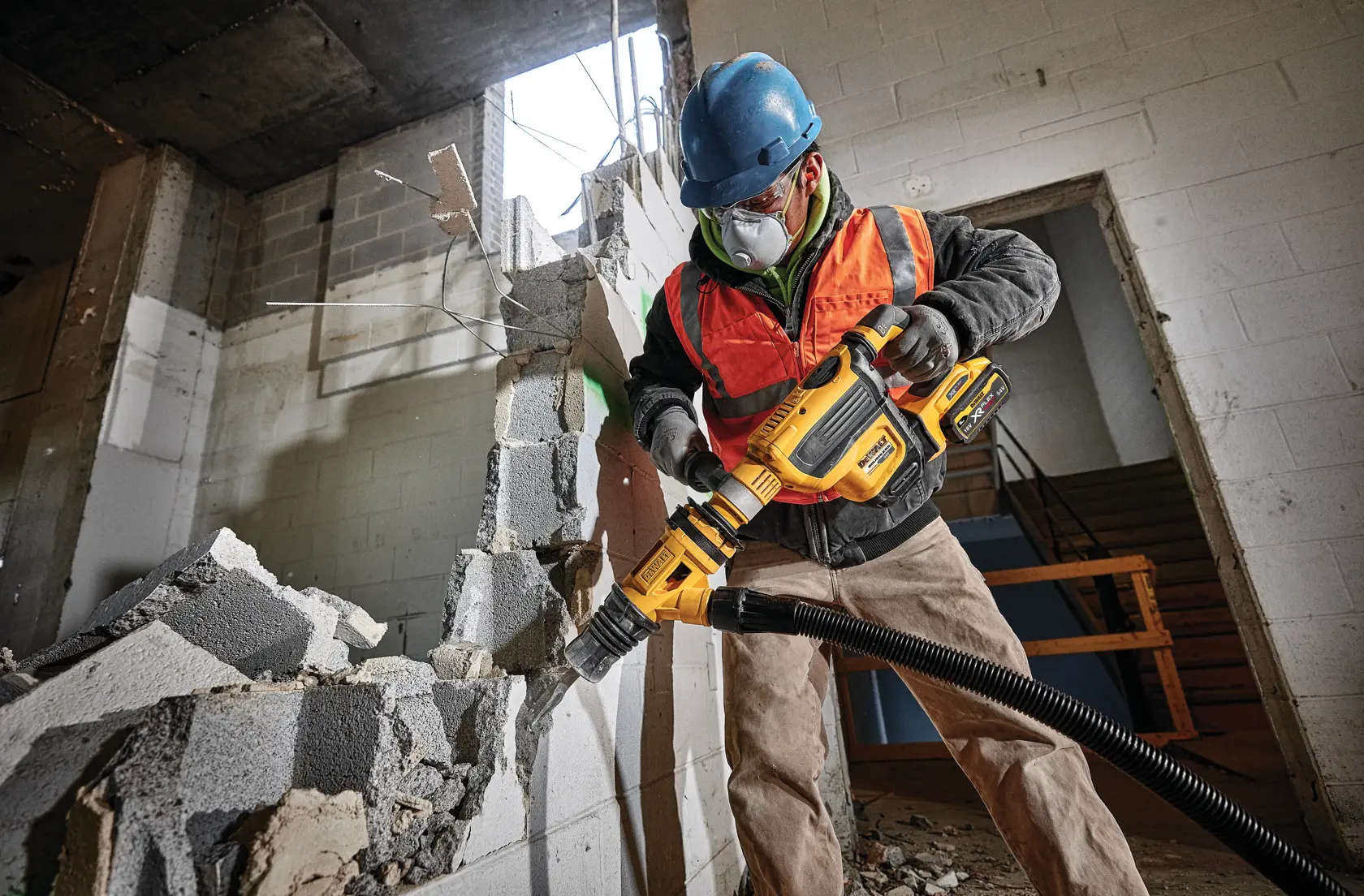 Worker uses a DEWALT power tool to demolish a concrete wall at a construction site while wearing safety gear.