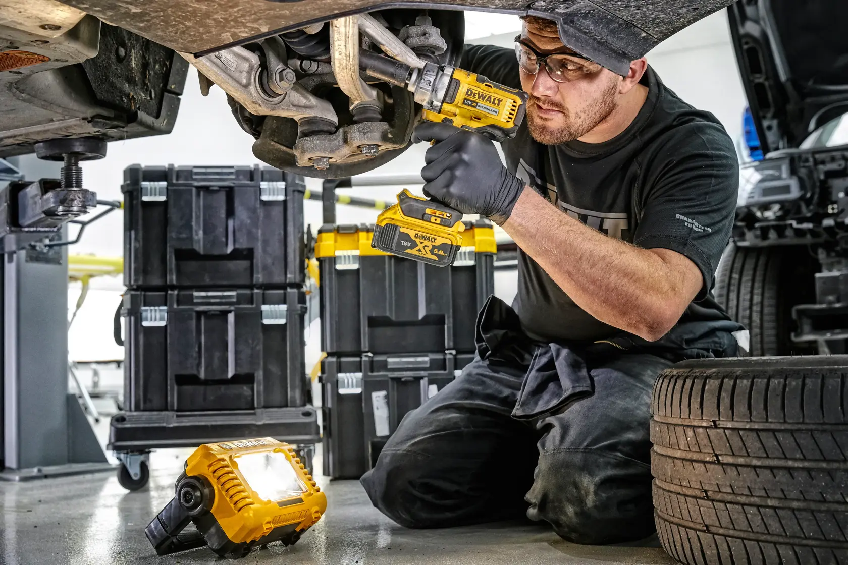 Persona usando herramientas DEWALT en la suspensión de un auto, luz de trabajo y cajas de almacenamiento cerca.