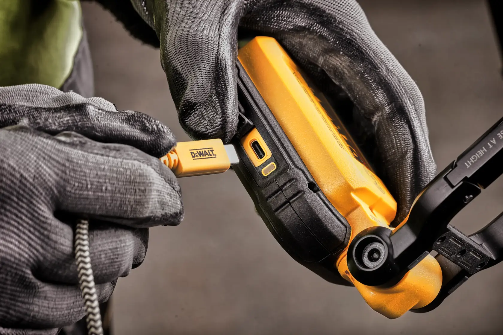 Nærbilde av hanskekledde hender som kobler en DEWALT USB-kabel til ladeporten på en gul DEWALT-enhet, sannsynligvis en bærbar arbeidslampe.
