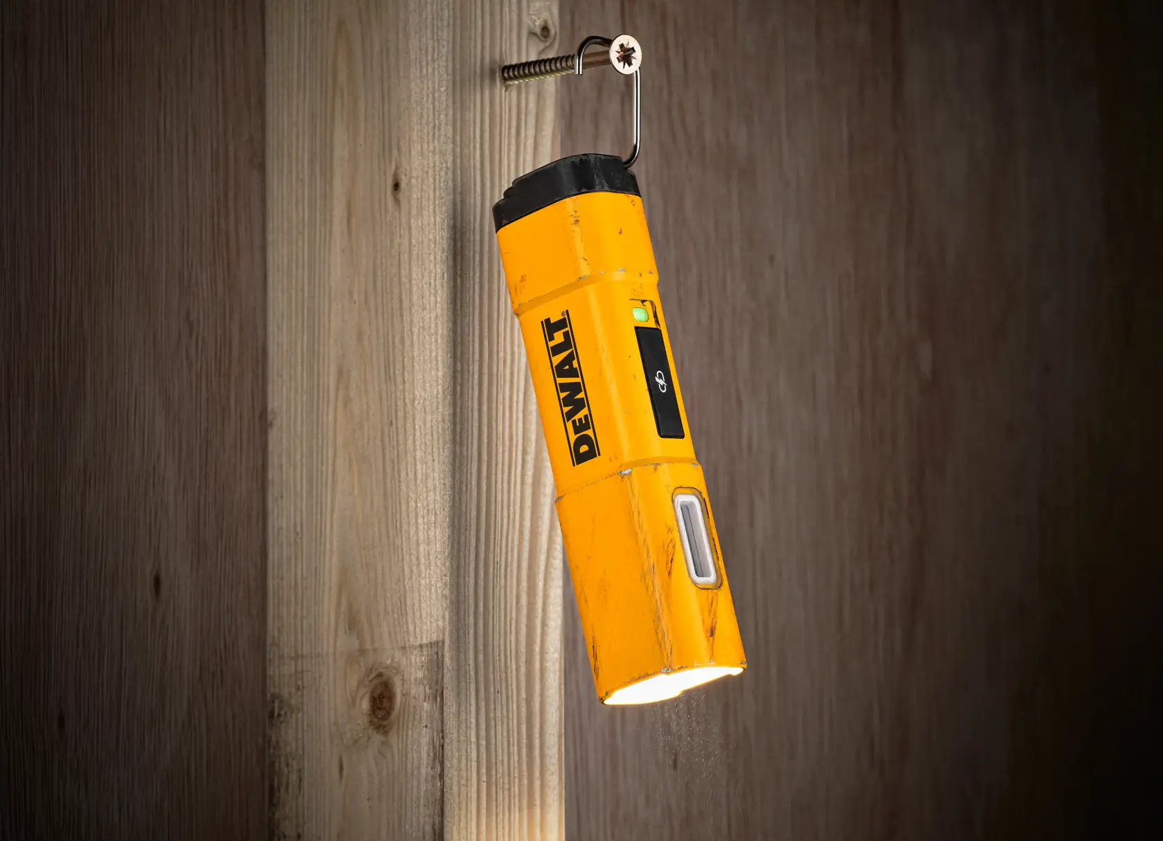 Et gult DEWALT DCL183 inspeksjonslys med svart topp, hengende fra en skrue på en trevegg og lyser nedover.