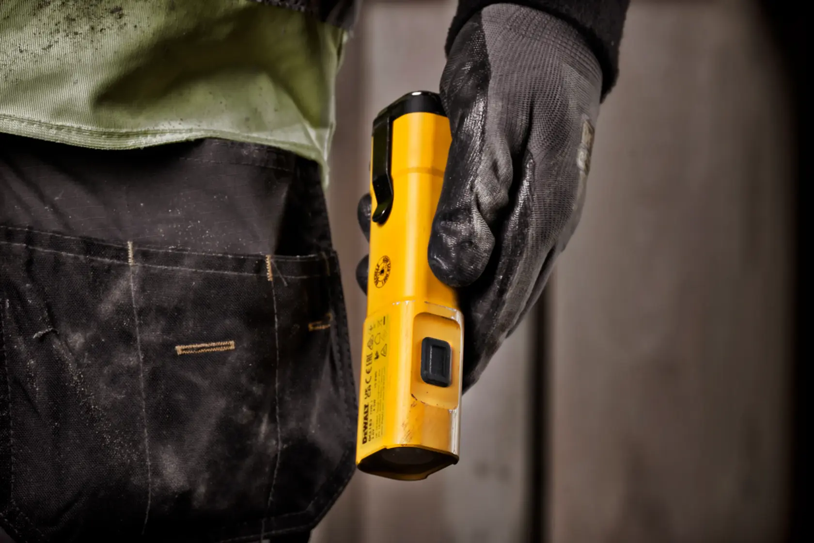 Nærbilde av en person med arbeidshanske som holder en gul DEWALT DCL183 bærbar arbeidslampe, stående ved siden av en verktøylomme.