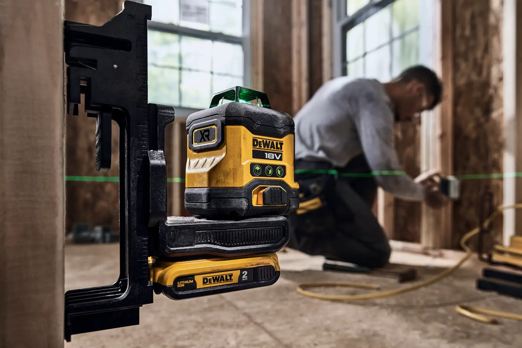 A DEWALT 18V XR green laser level tool is mounted on a clamp in a construction setting. A worker is in the background, kneeling and working on wooden framing, partially blurred for anonymity. The green laser line is visible across the workspace.