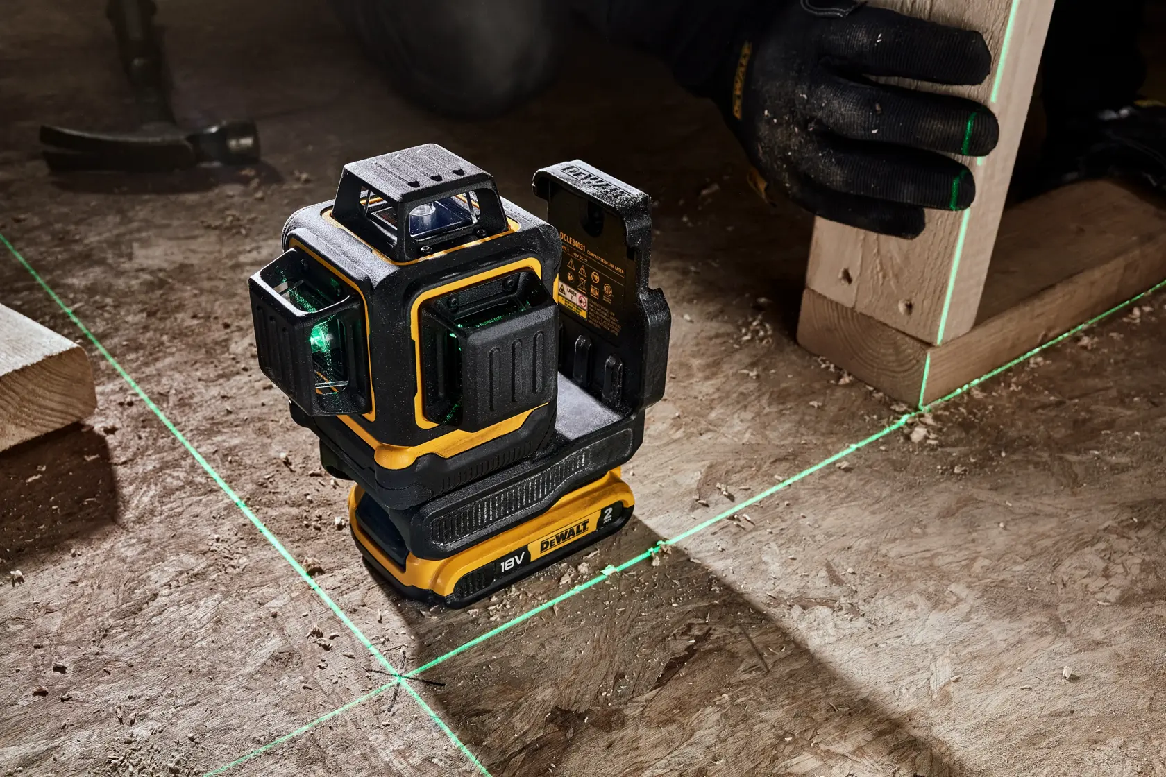 A DEWALT laser level tool projecting green laser lines on a wooden floor, used for accurate alignment during construction. A person wearing gloves is holding a wooden beam next to the laser lines.