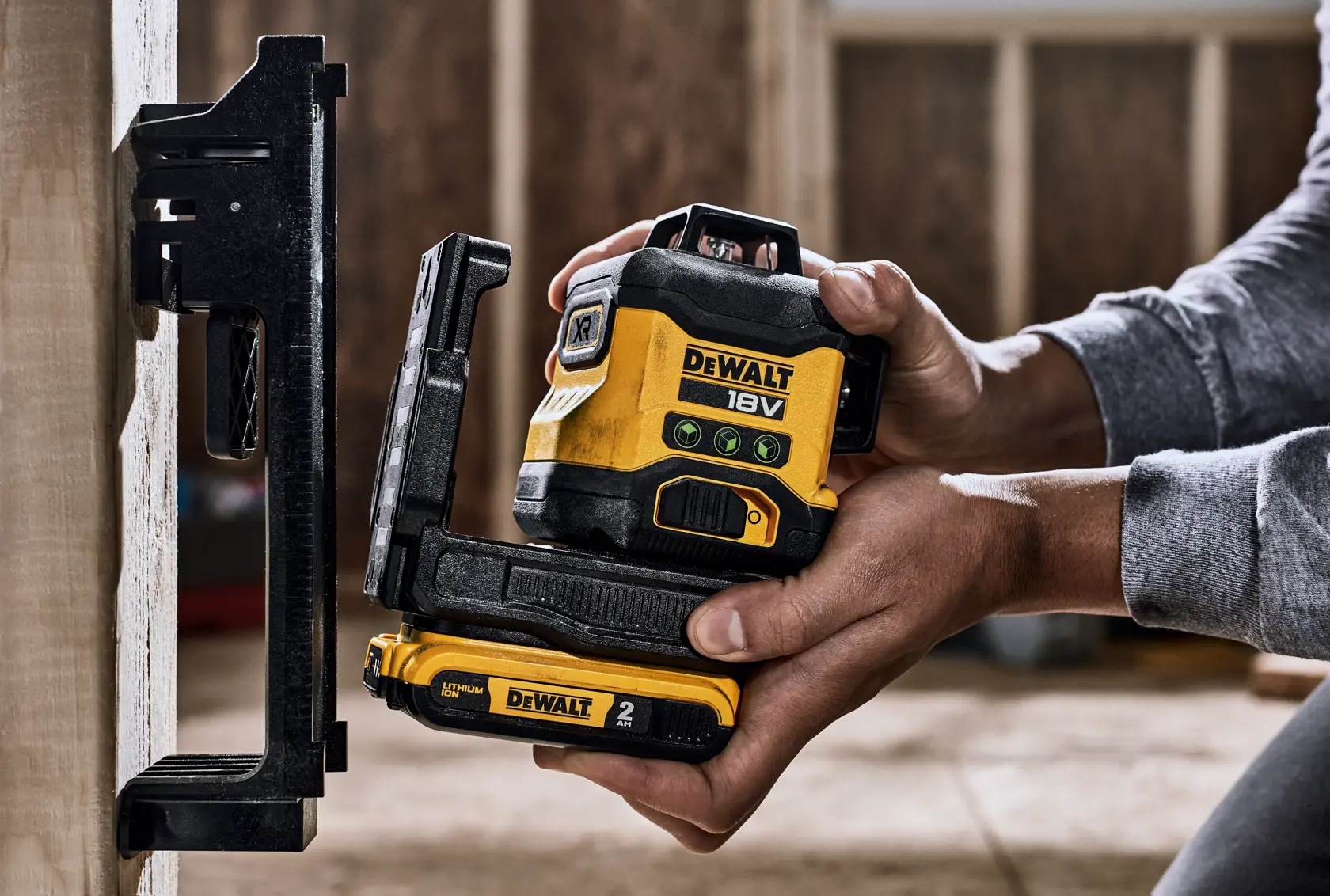 A close-up of hands positioning a DEWALT 18V laser level device, model DCLE34031N, against a vertical surface inside a construction site. The device is yellow and black with clear labeling and battery compartment visible.