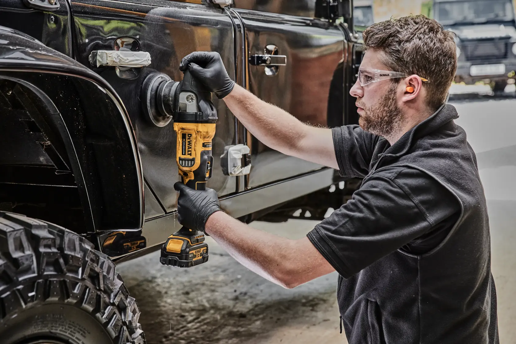 Eine Person mit schwarzen Handschuhen verwendet eine kabellose DEWALT-Poliermaschine (Modell DCM848N), um die Seite eines schwarzen Geländewagens zu polieren. Die Szene findet in einer Werkstatt oder Garage statt.