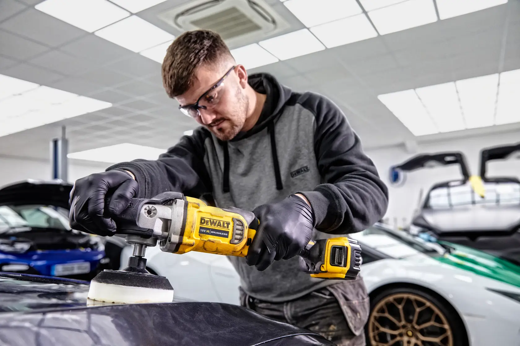 Person using DEWALT DCM849P2 polisher on car surface in a modern garage with luxury vehicles in the background.