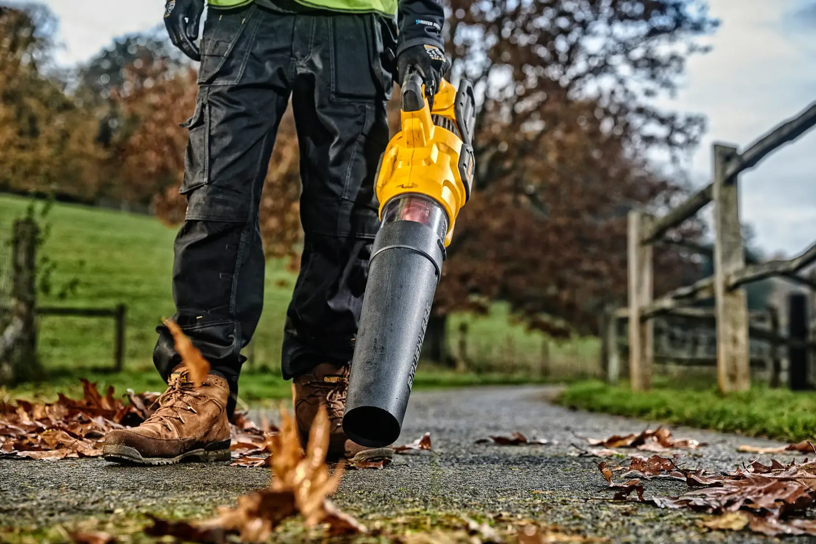 Person using a yellow DEWALT leaf blower on a paved path with scattered autumn leaves outdoors.
