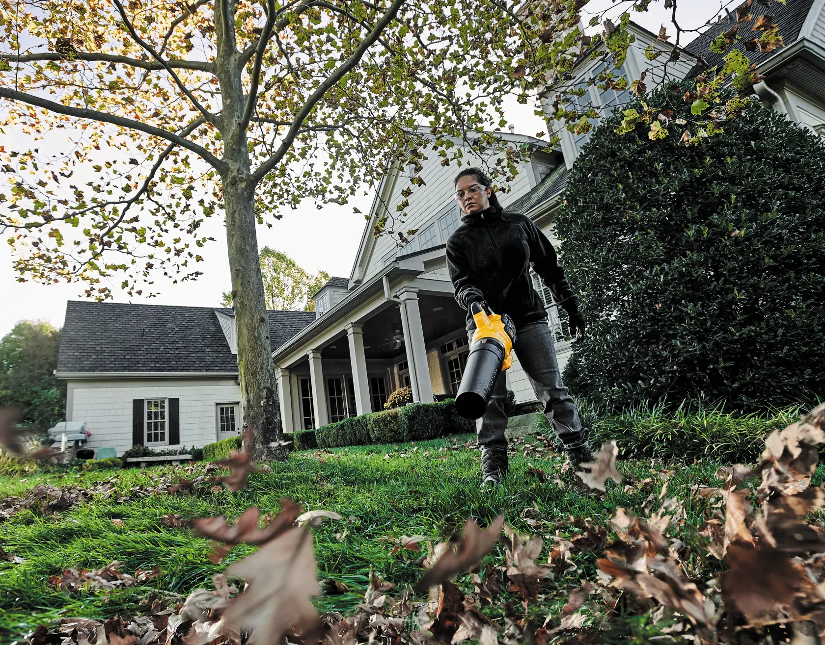 Person uses a DEWALT leaf blower to clear leaves in a yard outside a house.