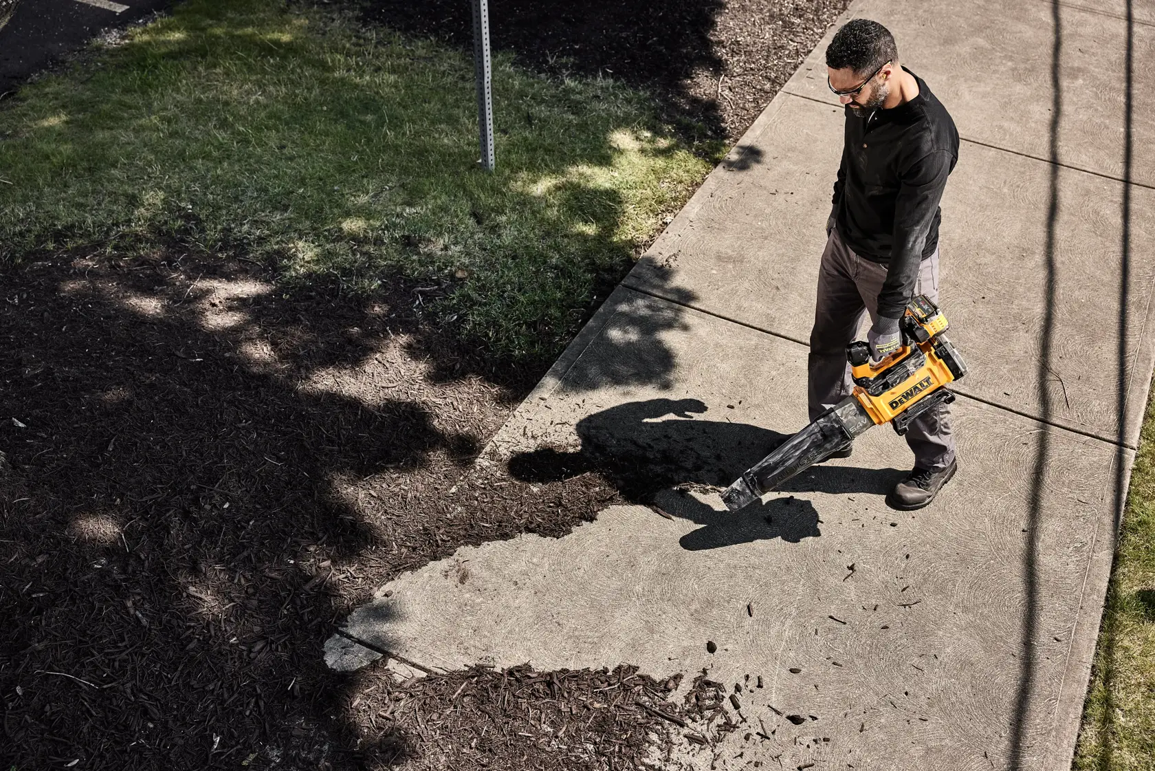 Person uses a DEWALT cordless leaf blower to clear debris from a sidewalk near grass and mulch.