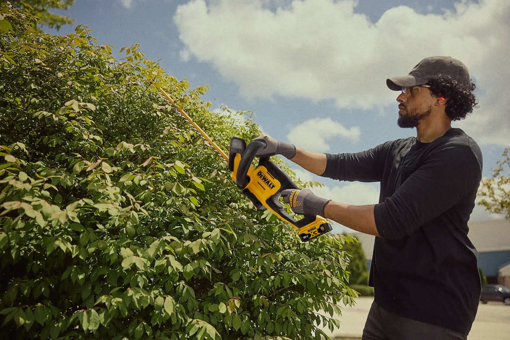 Person schneidet Hecken mit einem kabellosen DEWALT-Trimmer bei Sonnenschein.